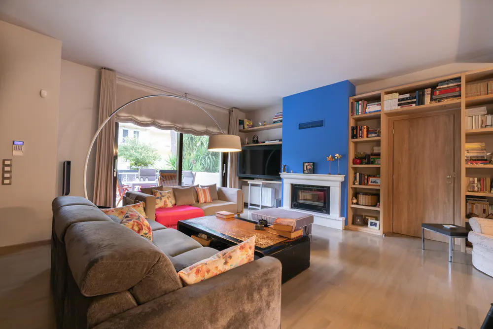 Living room with a gray sectional sofa, a blue accent wall with a fireplace, and built-in bookshelves. A large window overlooks an outdoor patio.
