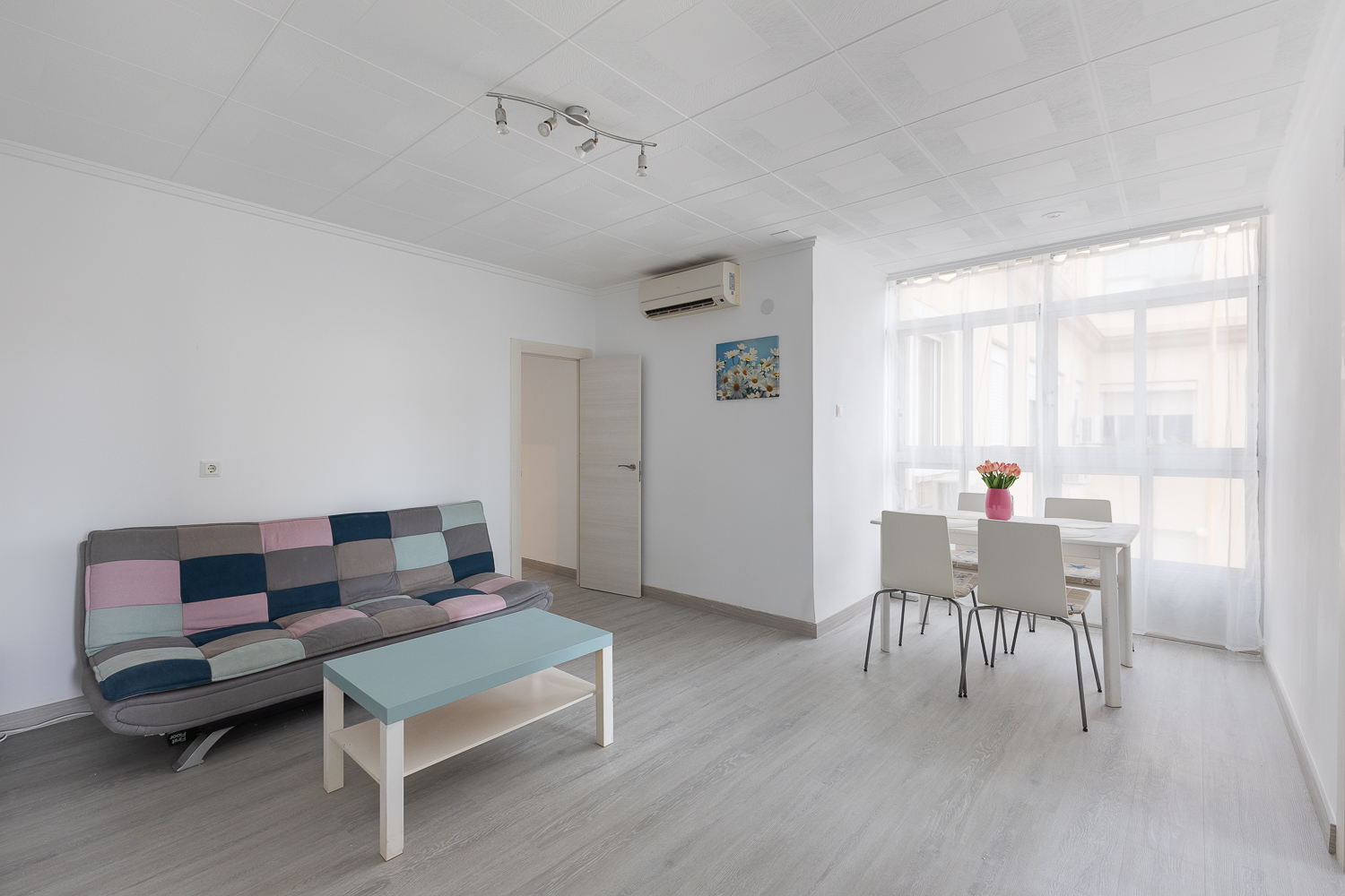 Bright living room with a multicolored patchwork sofa, light blue coffee table, and a dining area with four white chairs. [AI-generated text]