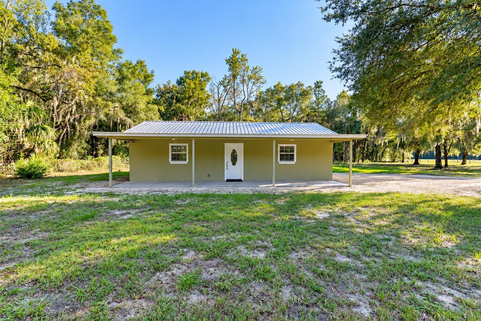 Single-story olive green house with a white metal roof and front porch, surrounded by trees on a grassy lot. [AI-generated text]