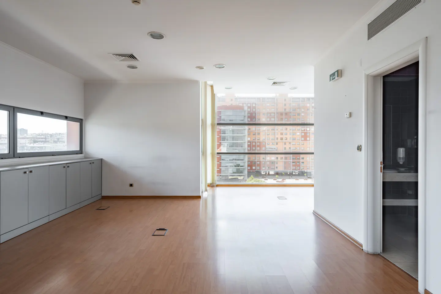 Bright, empty office space with wood floors, white walls, and large windows overlooking a city. A doorway leads to a bathroom.
