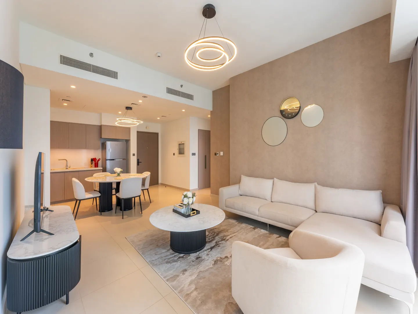 Open-concept living space with a white sofa, round marble coffee table, and dining area with modern lighting. Neutral color palette.