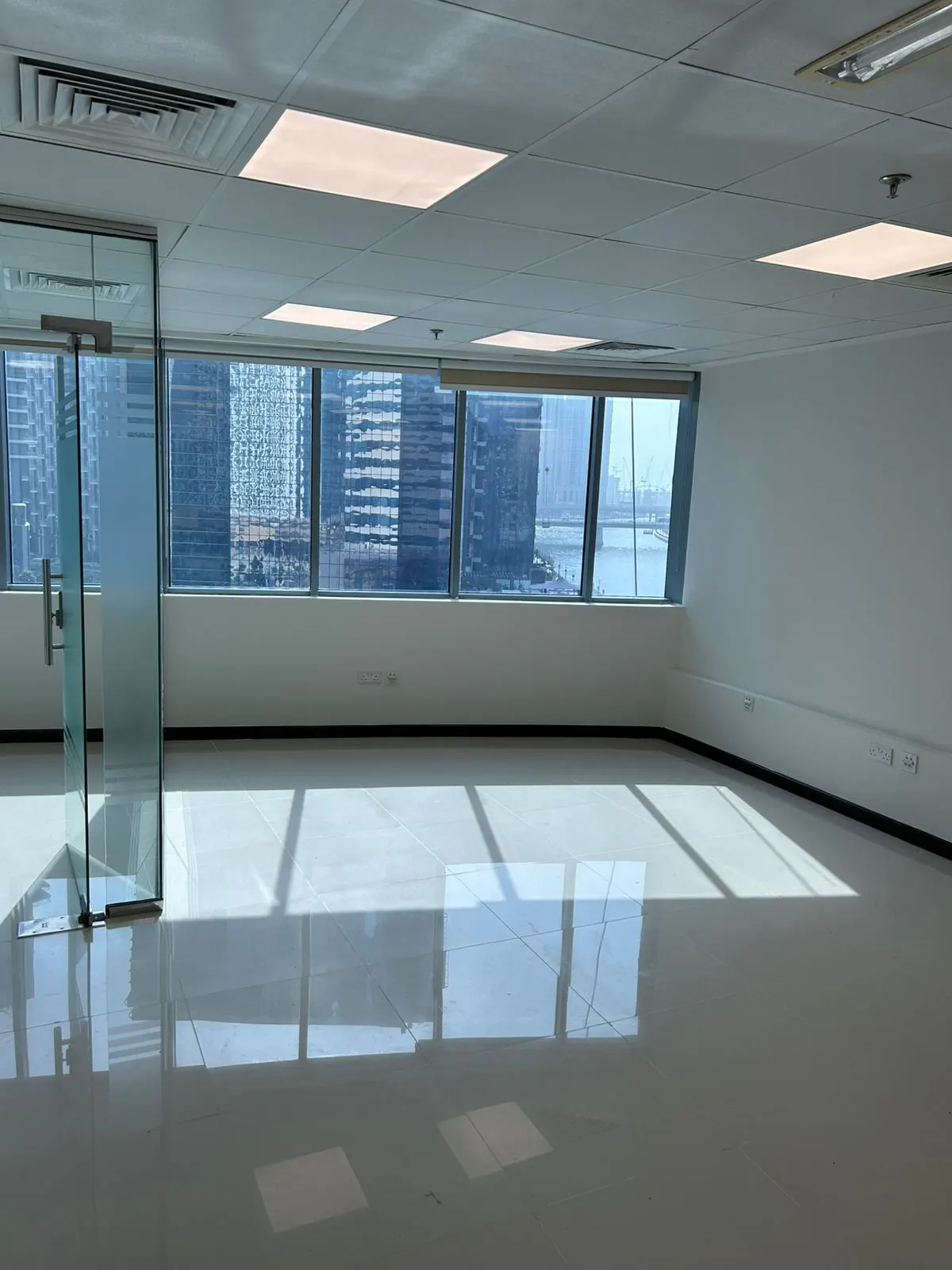 Empty office space with white walls, tile floor, and large windows overlooking city buildings and water. A glass door is on the left.