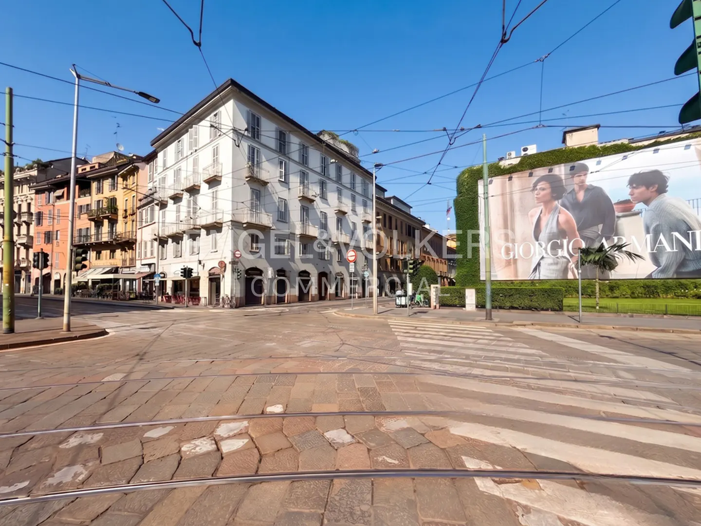 A sunny street corner in Milan with cobblestone roads, tram tracks, historic buildings, and a large Giorgio Armani billboard.