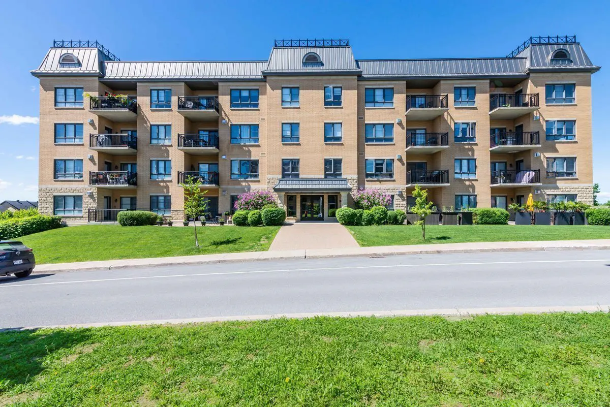 A four-story tan brick apartment building with black balconies and a gray roof under a blue sky.
