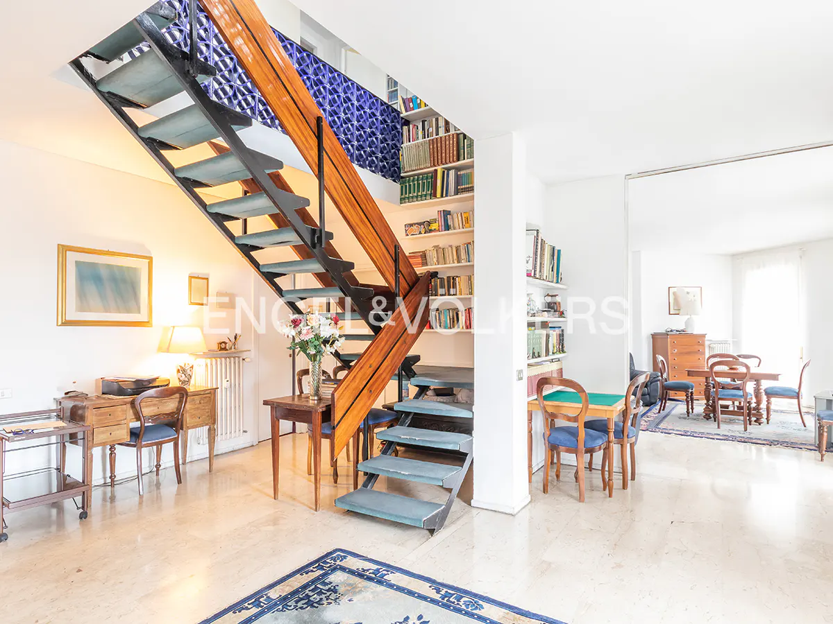 Bright, open living room with a staircase, bookshelves, and marble floors. The staircase has wooden railings and blue steps. A blue rug is on the floor.