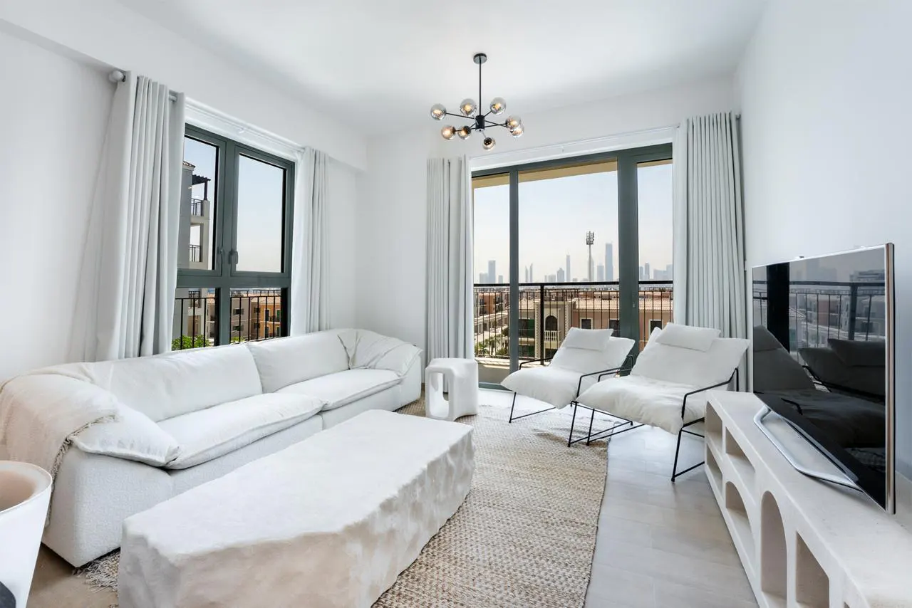 Bright living room with white sofa, chairs, and textured coffee table. Large windows offer a city view. Modern chandelier overhead.