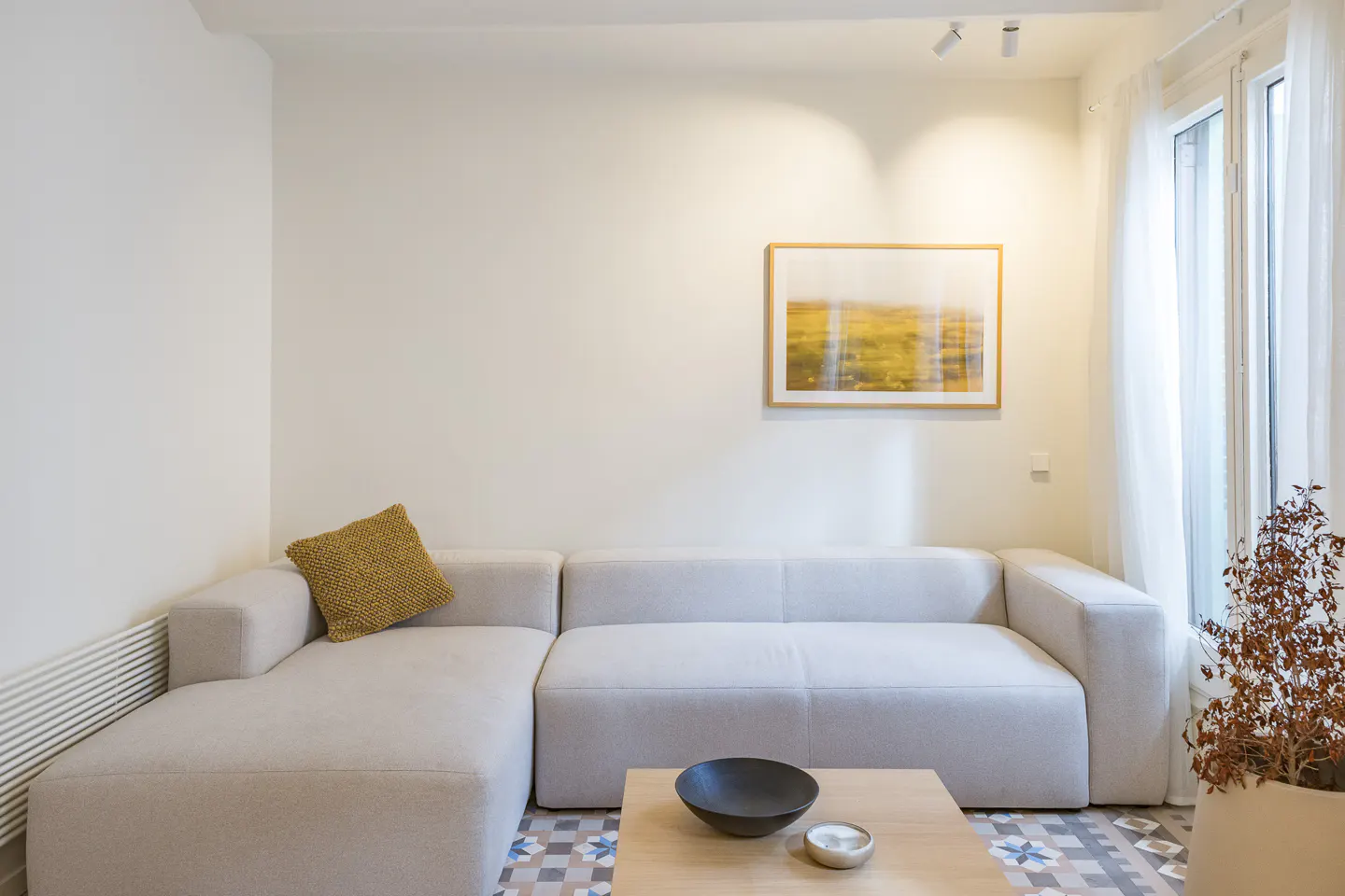 Living room with a white sectional sofa, a wooden coffee table, and a framed abstract art piece on the wall. A window with sheer curtains is on the right.