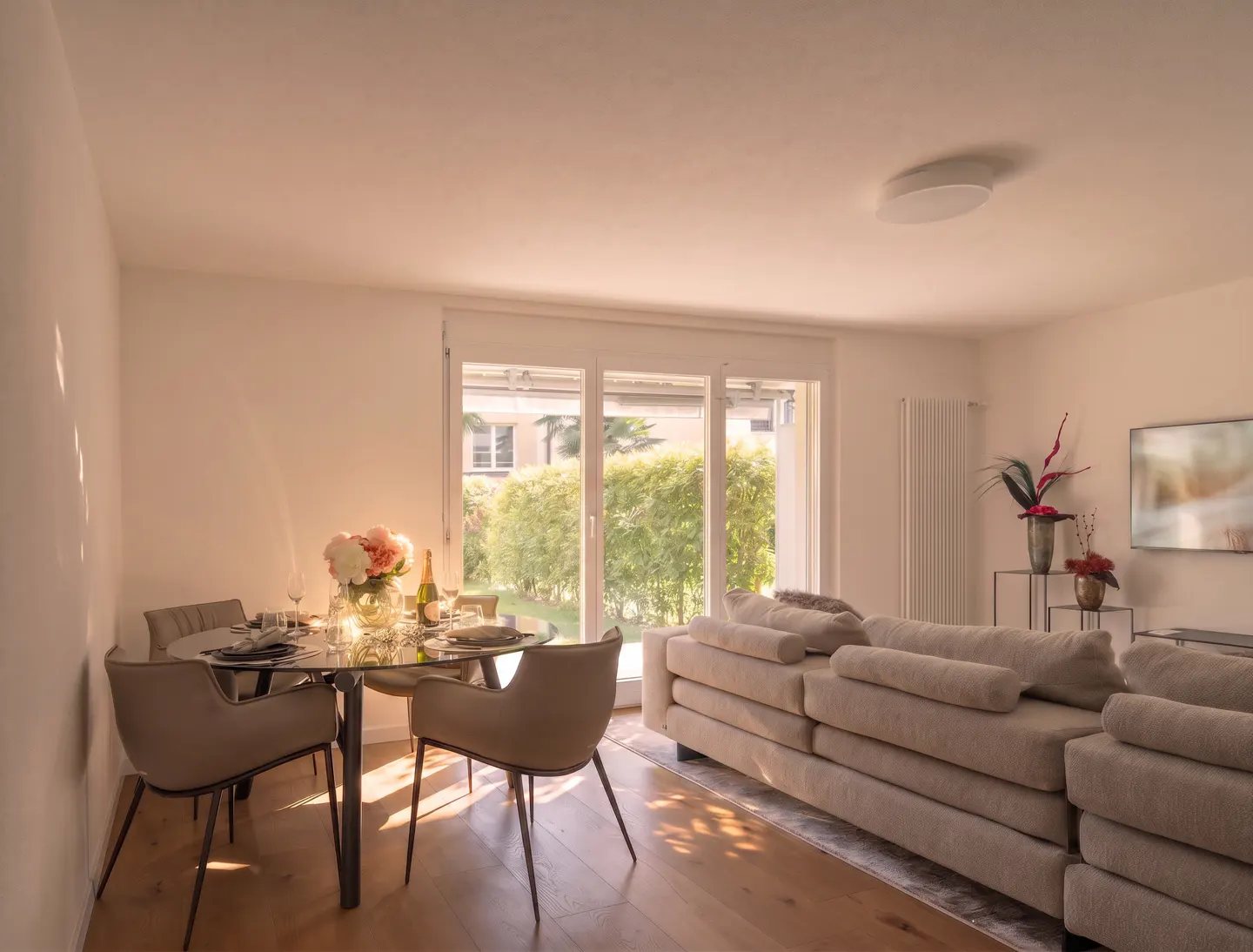 Bright living room with a beige sofa, glass dining table, four chairs, and large glass doors opening to a green garden.