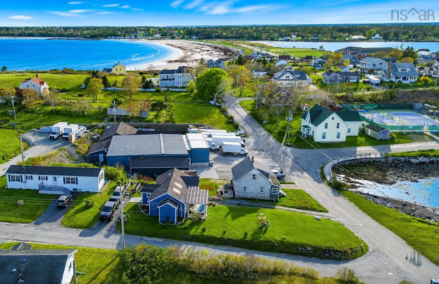 Aerial view of a coastal town with houses, green lawns, a sandy beach, and blue ocean under a partly cloudy sky.