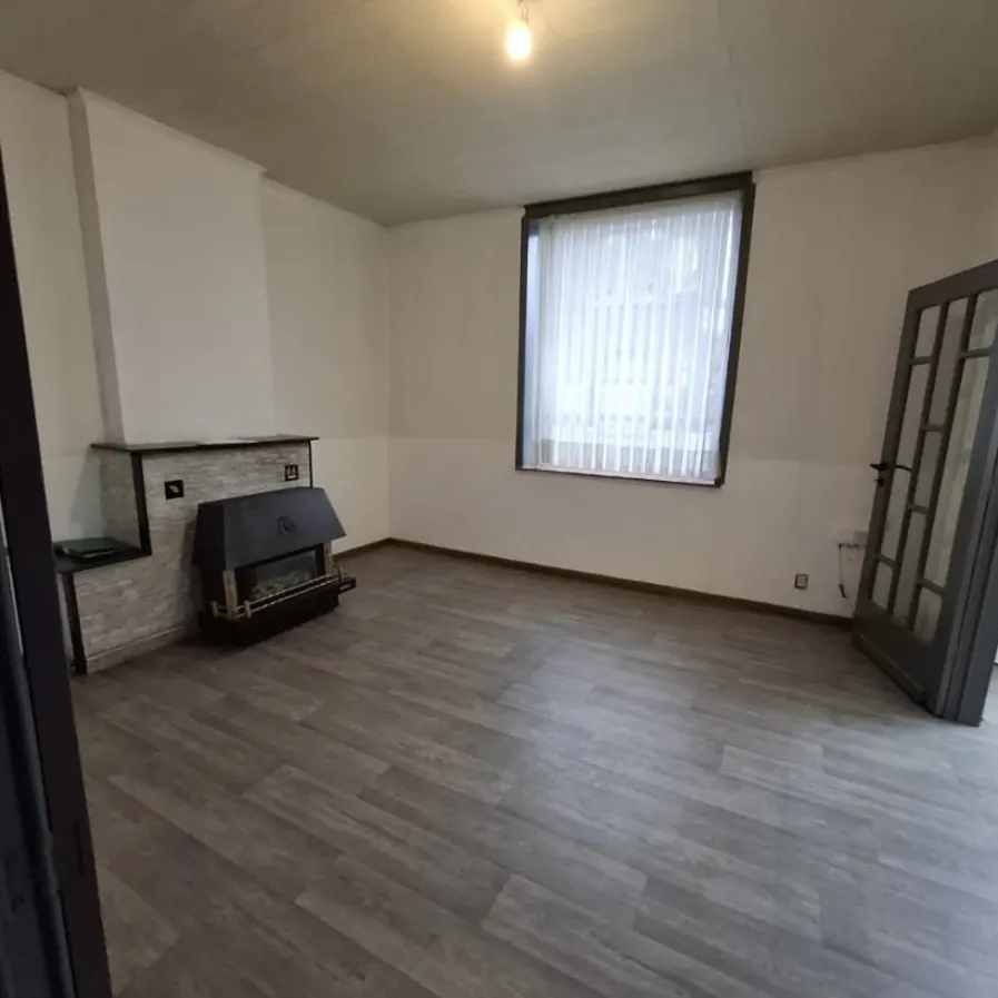 Empty room with gray wood floors, white walls, a fireplace, a window with white curtains, and a gray door.