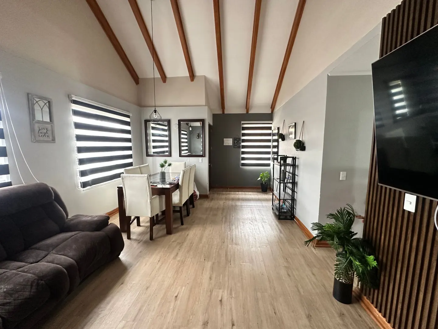 A living and dining area with light wood floors, a brown sofa, a dining table with white chairs, and a TV on a wood-paneled wall.