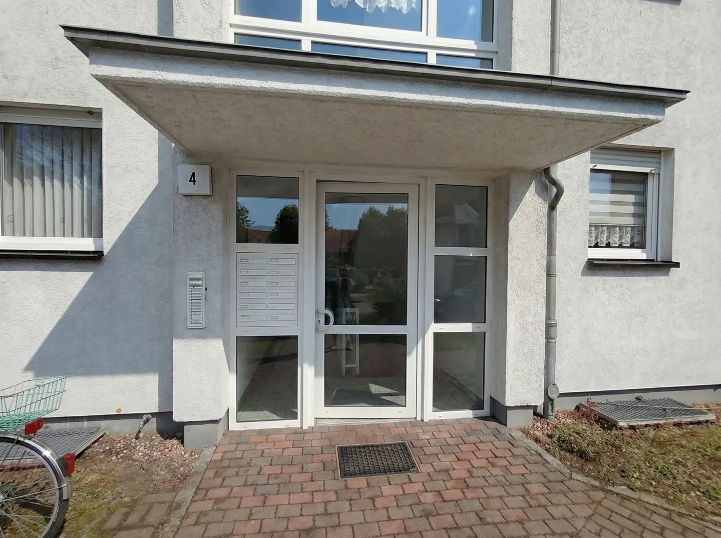 Apartment building entrance with white door, mailbox, and number 4. A brick path leads to the door. A bicycle is parked nearby.
