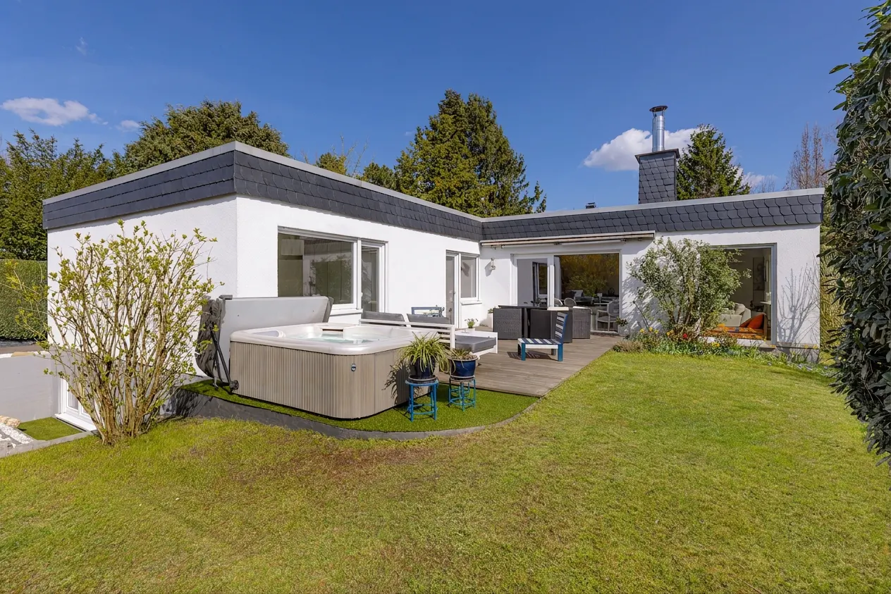 Exterior view of a modern white house with a gray roof, a wooden deck, a hot tub, and a green lawn.