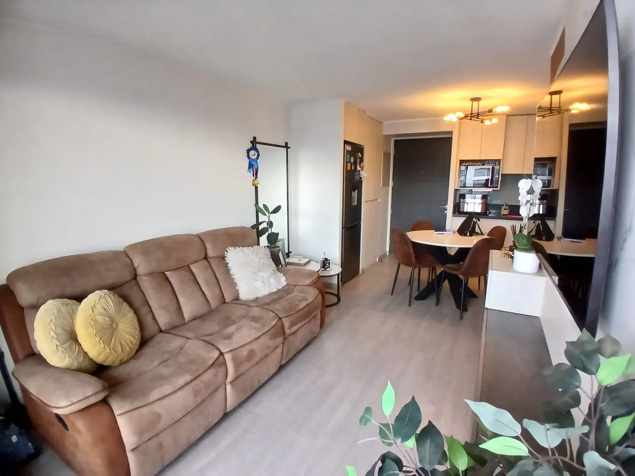 Living room with a brown sofa, round pillows, and a white fluffy pillow. A dining table with chairs is in the background.