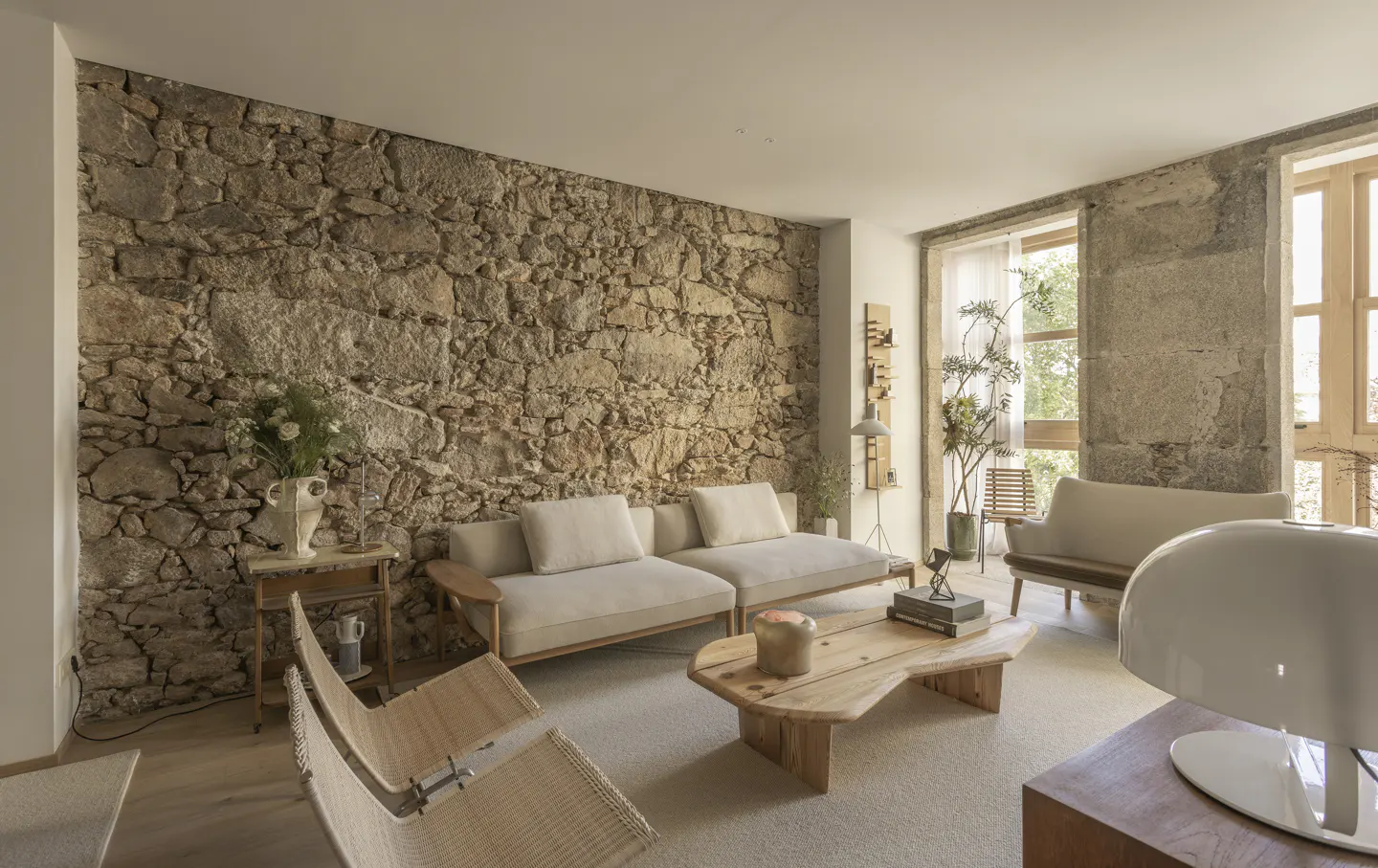 Living room with stone wall, beige sofas, and wooden coffee table. Large windows let in natural light. Neutral color palette.