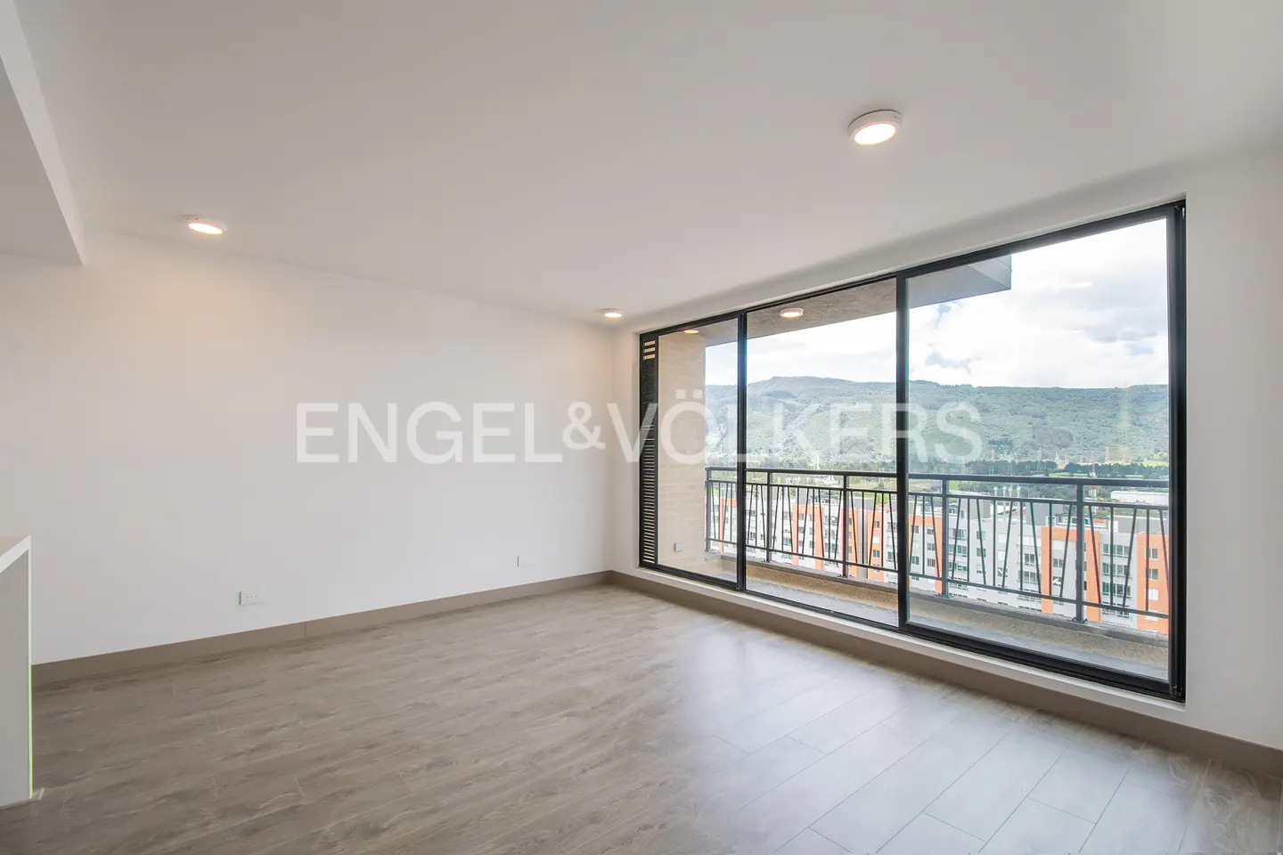 Bright, empty room with light wood floors and white walls. Large windows offer a view of mountains and buildings. Engel & Völkers logo visible.