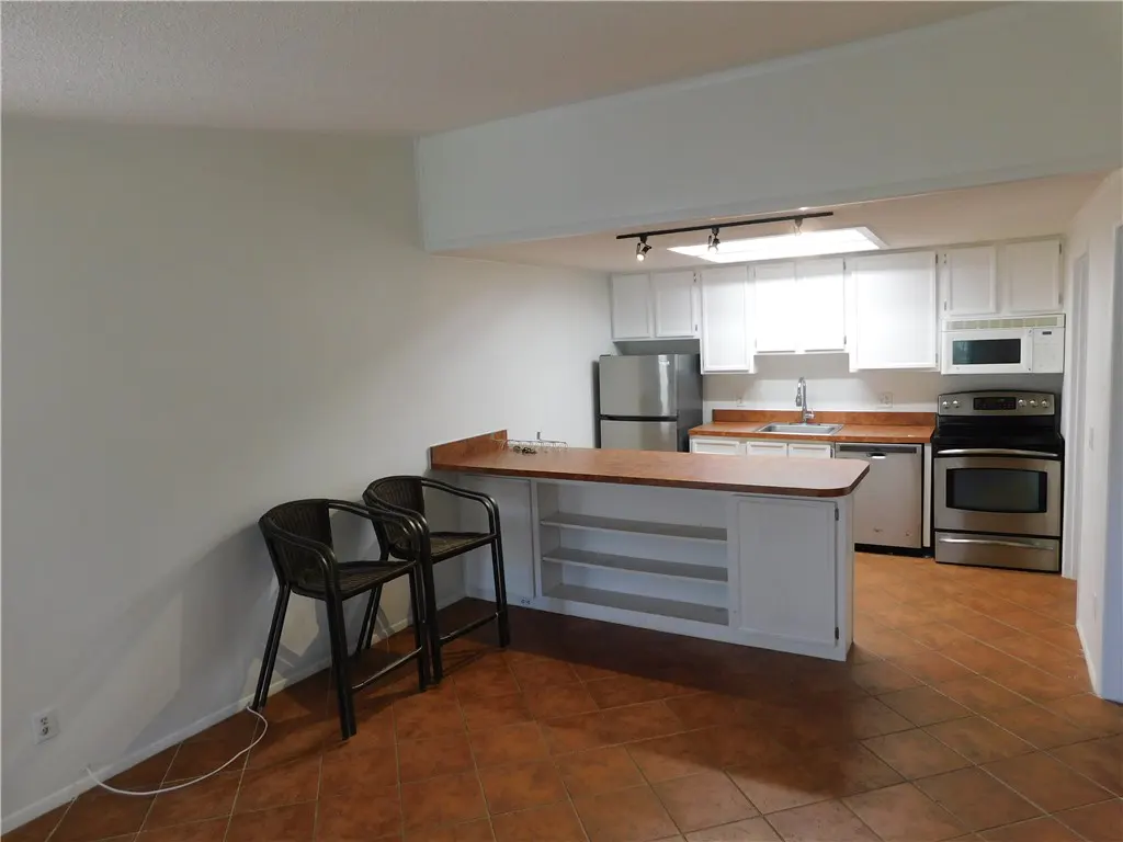 A kitchen with white cabinets, stainless steel appliances, and a brown tile floor. Two black chairs sit at a counter.