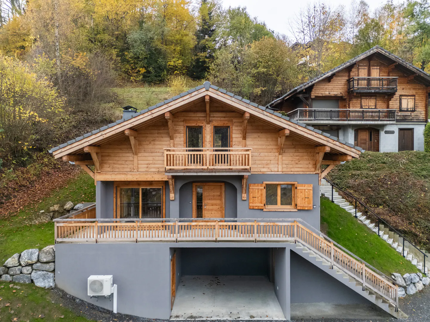 Two-story chalet with a gray base and wooden upper levels, set on a hillside with autumn foliage. Balconies and a garage are visible.