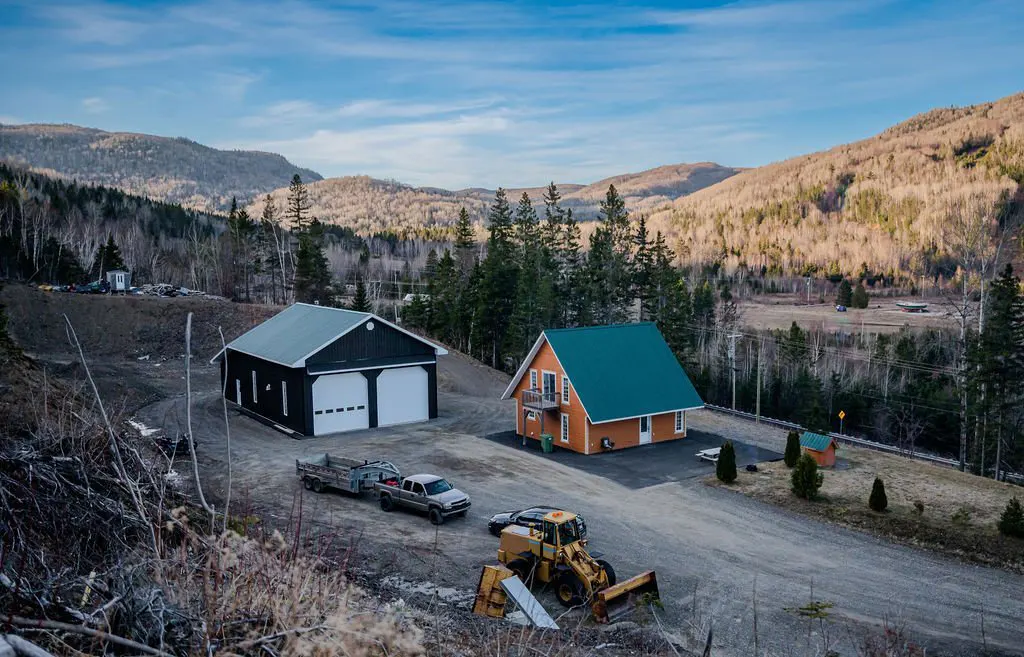 A rural property with an orange house, green roof, black garage, and construction equipment, set against a mountain backdrop.