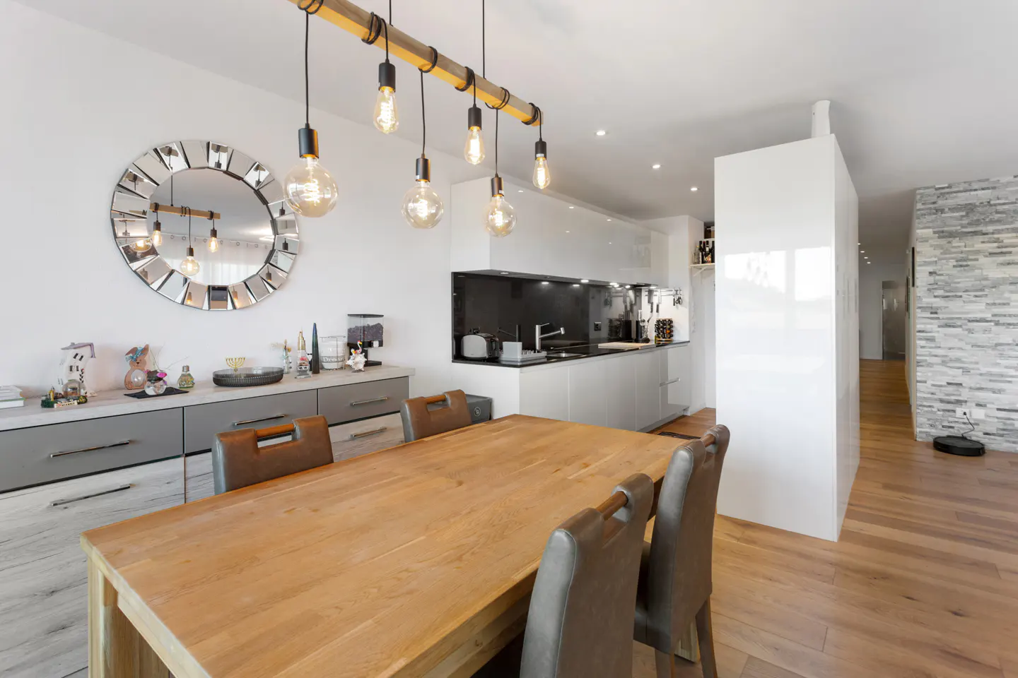 Bright, modern dining area with a wooden table, leather chairs, and a unique light fixture. White kitchen and stone wall in the background.