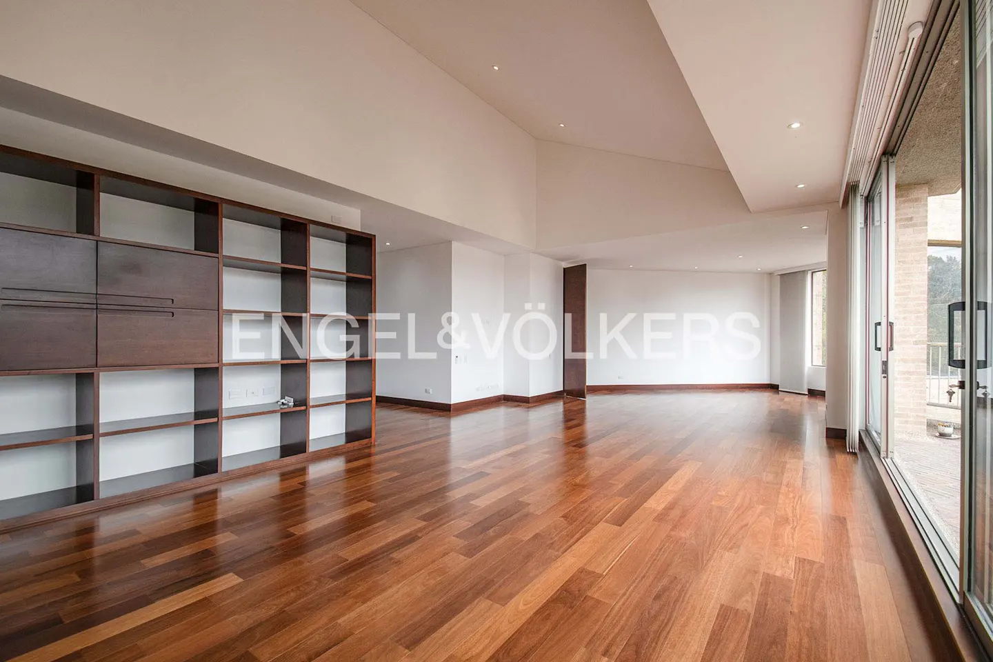 Spacious living room with hardwood floors, a large dark wood bookshelf, and sliding glass doors to a balcony. White walls and ceiling.