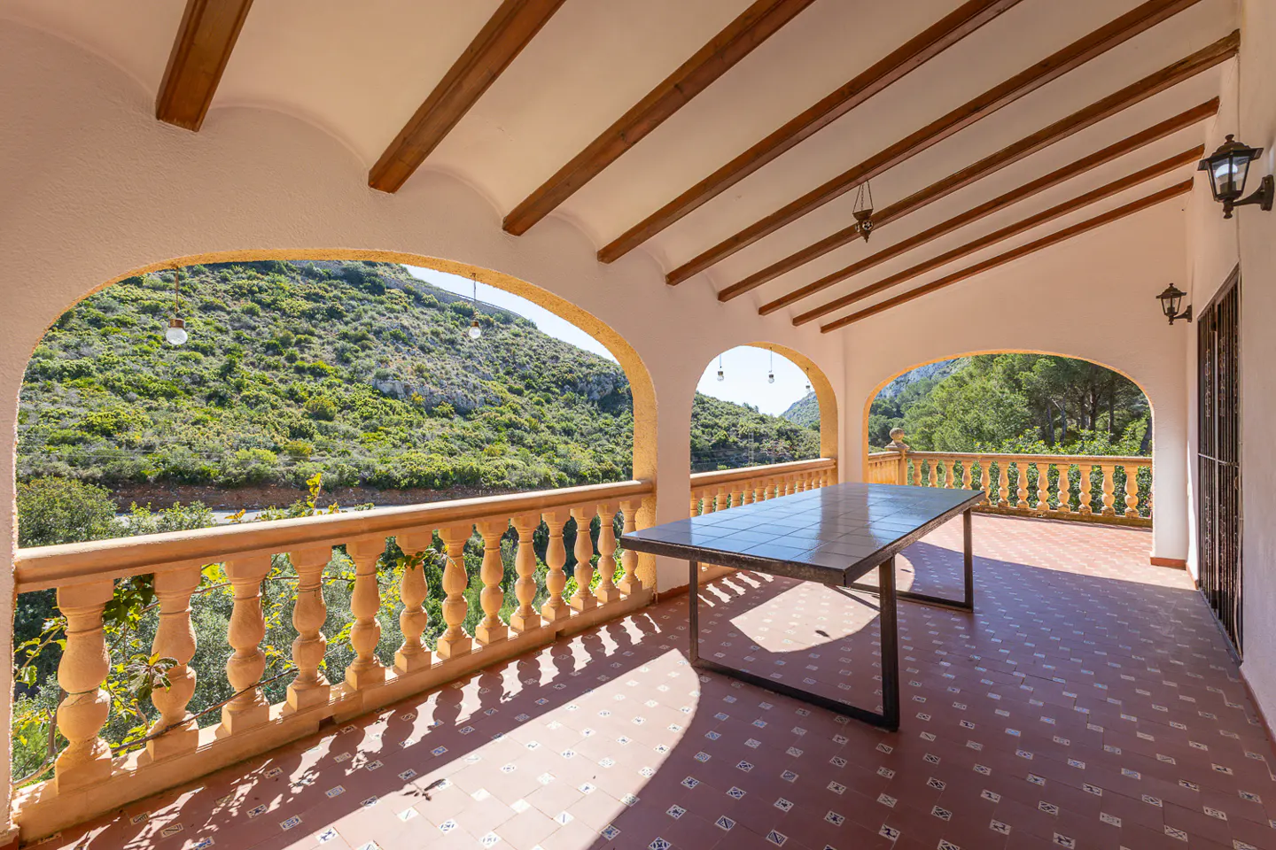 Covered patio with arches, a stone balustrade, and a tiled floor. A table sits in the center, with a green hillside visible through the arches.