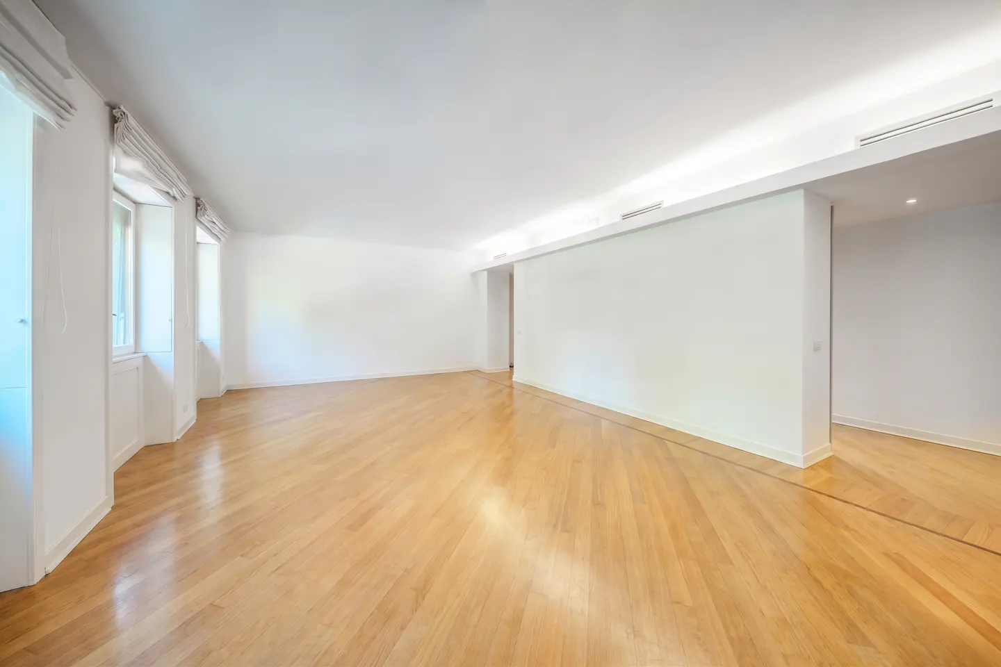 Spacious, empty room with white walls, polished light wood floors, and large windows with white curtains.