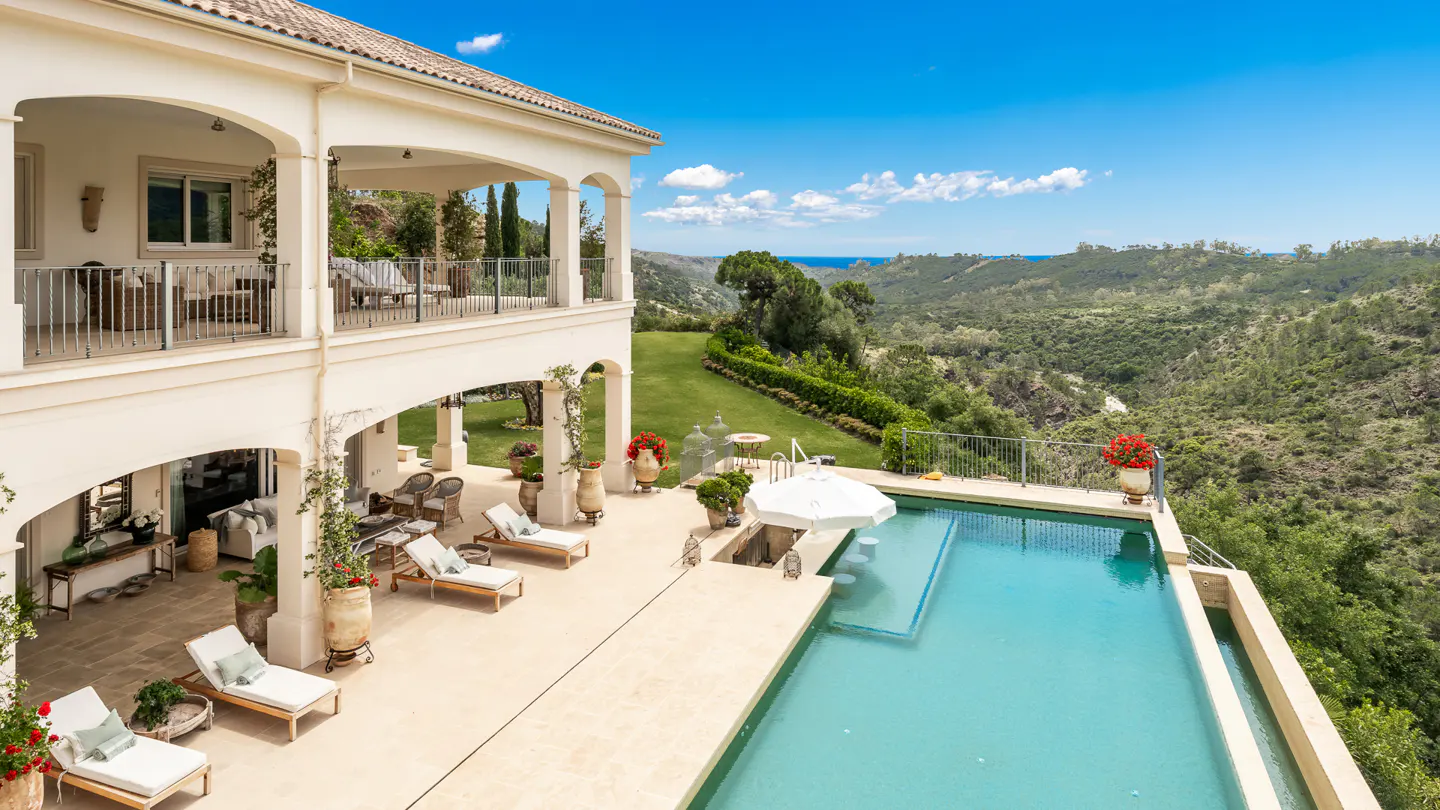 Luxury villa with pool overlooking green hills. White lounge chairs line the patio. Blue sky.