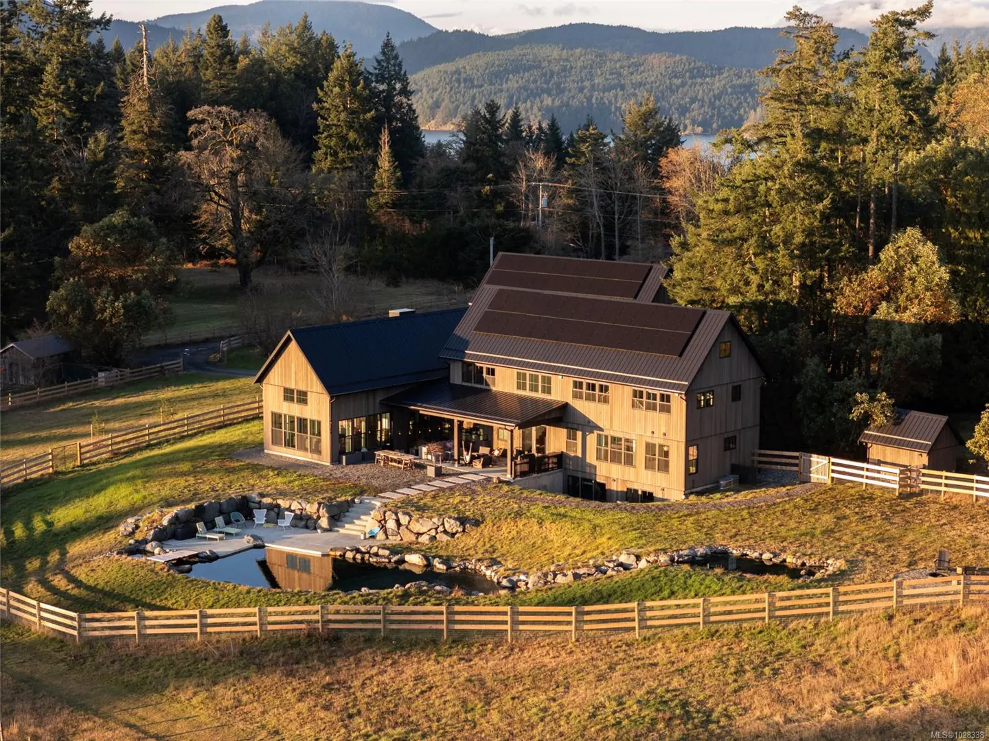 Aerial view of a modern farmhouse with a dark roof, a pond, and a wooden fence in a grassy field. Trees and mountains are in the background.