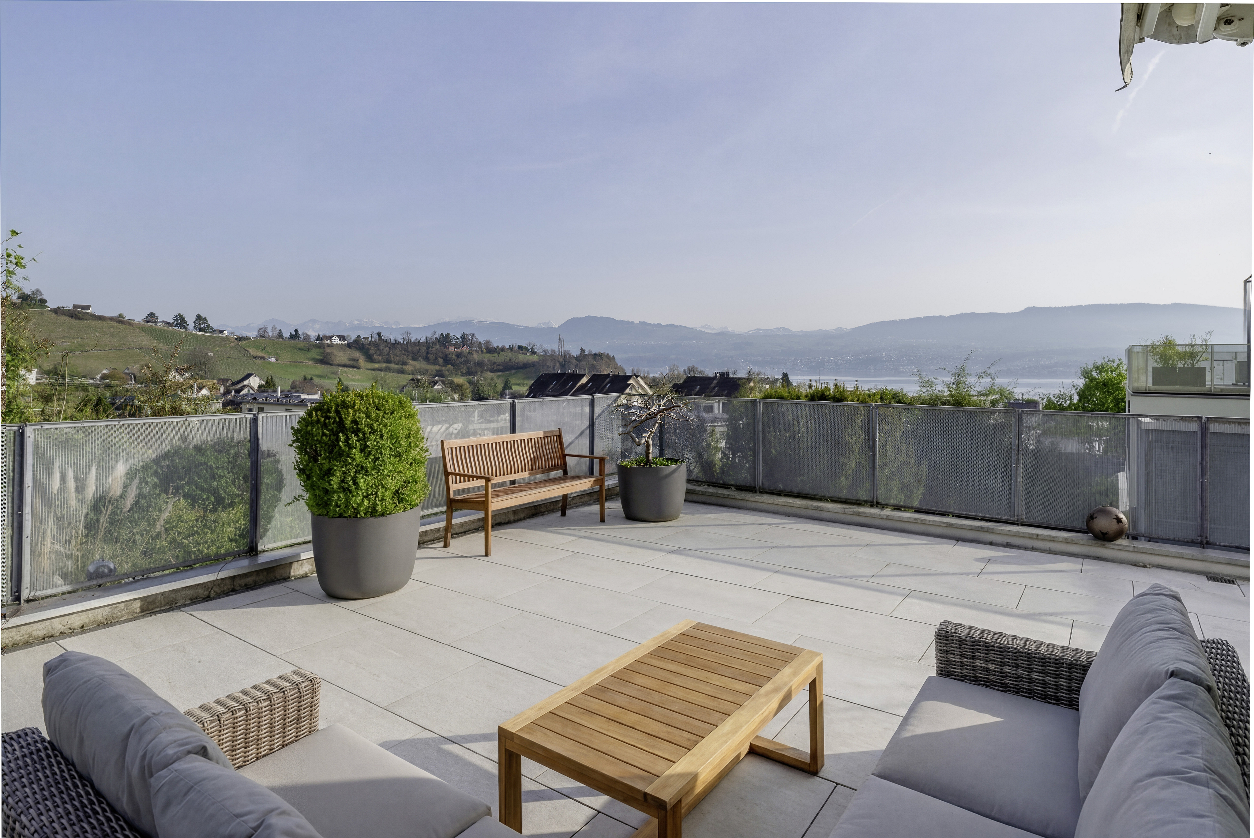 A rooftop patio with a wooden bench, table, and gray sofas overlooks a scenic view of rolling hills and a lake.