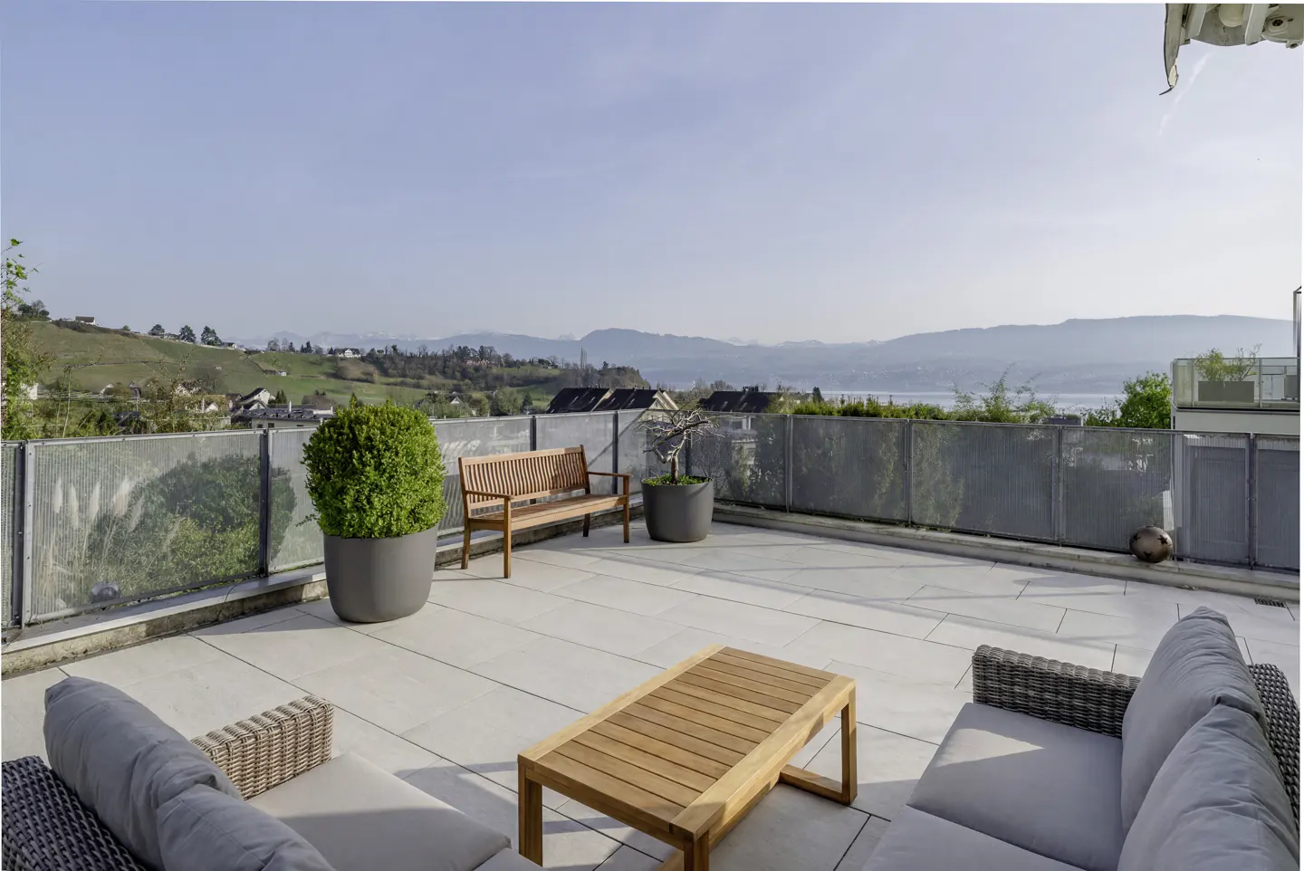 A rooftop patio with a wooden bench, table, and gray sofas overlooks a scenic view of rolling hills and a lake.