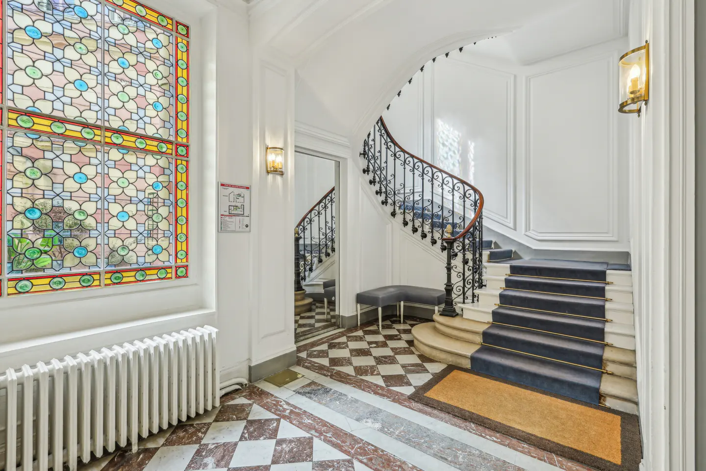 Elegant foyer with a stained-glass window, checkered marble floor, and a curved staircase with a dark blue runner.