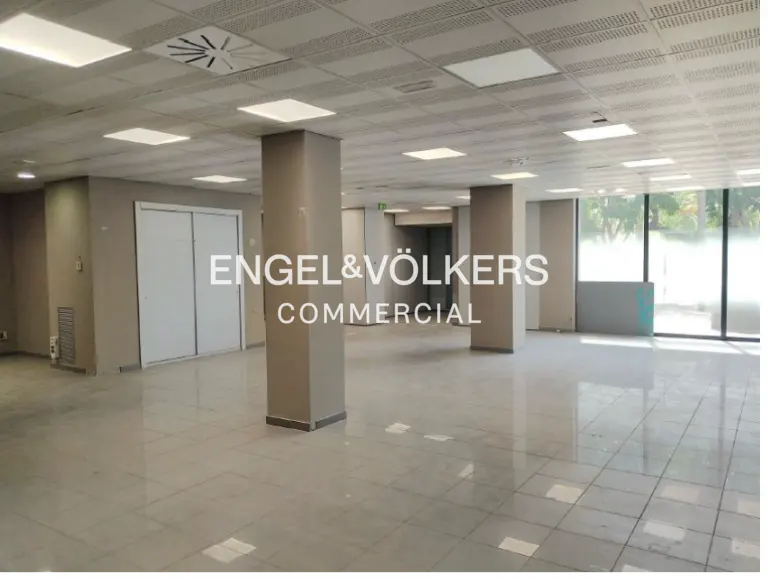 Empty commercial space with gray tile floor, columns, and white ceiling panels with square lights. Large windows let in natural light.