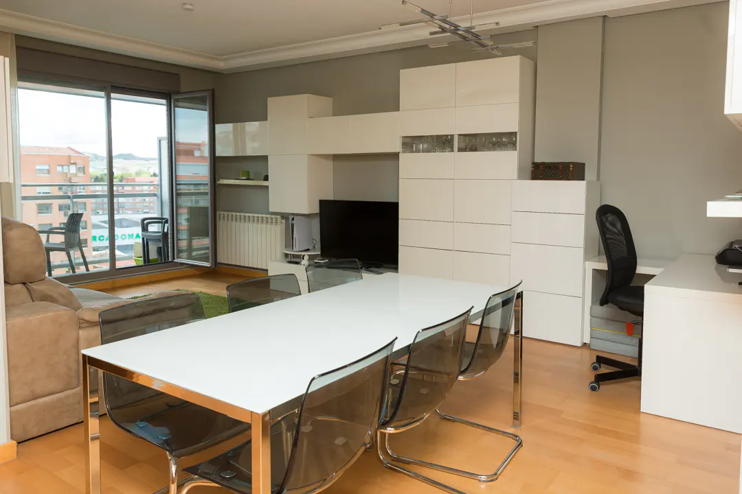 Living room with a white table and transparent chairs, a beige sofa, and a balcony with a view of buildings.