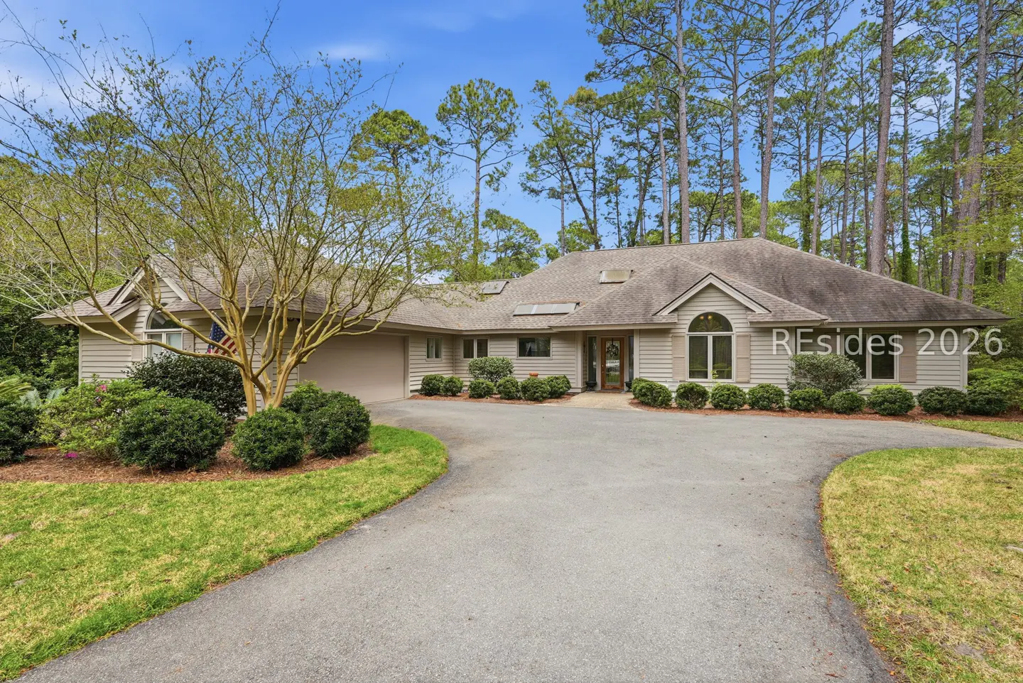 Beige single-story house with a gray roof, surrounded by trees and green bushes, with a gray driveway.
