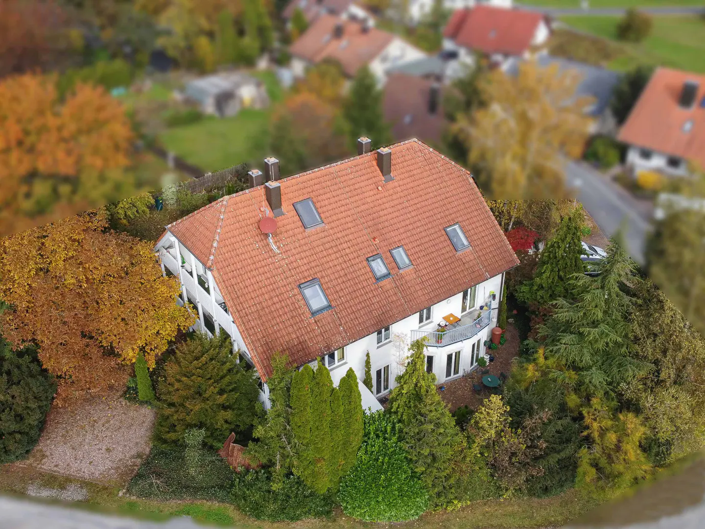 Aerial view of a white house with a red tile roof, surrounded by green and autumn-colored trees.