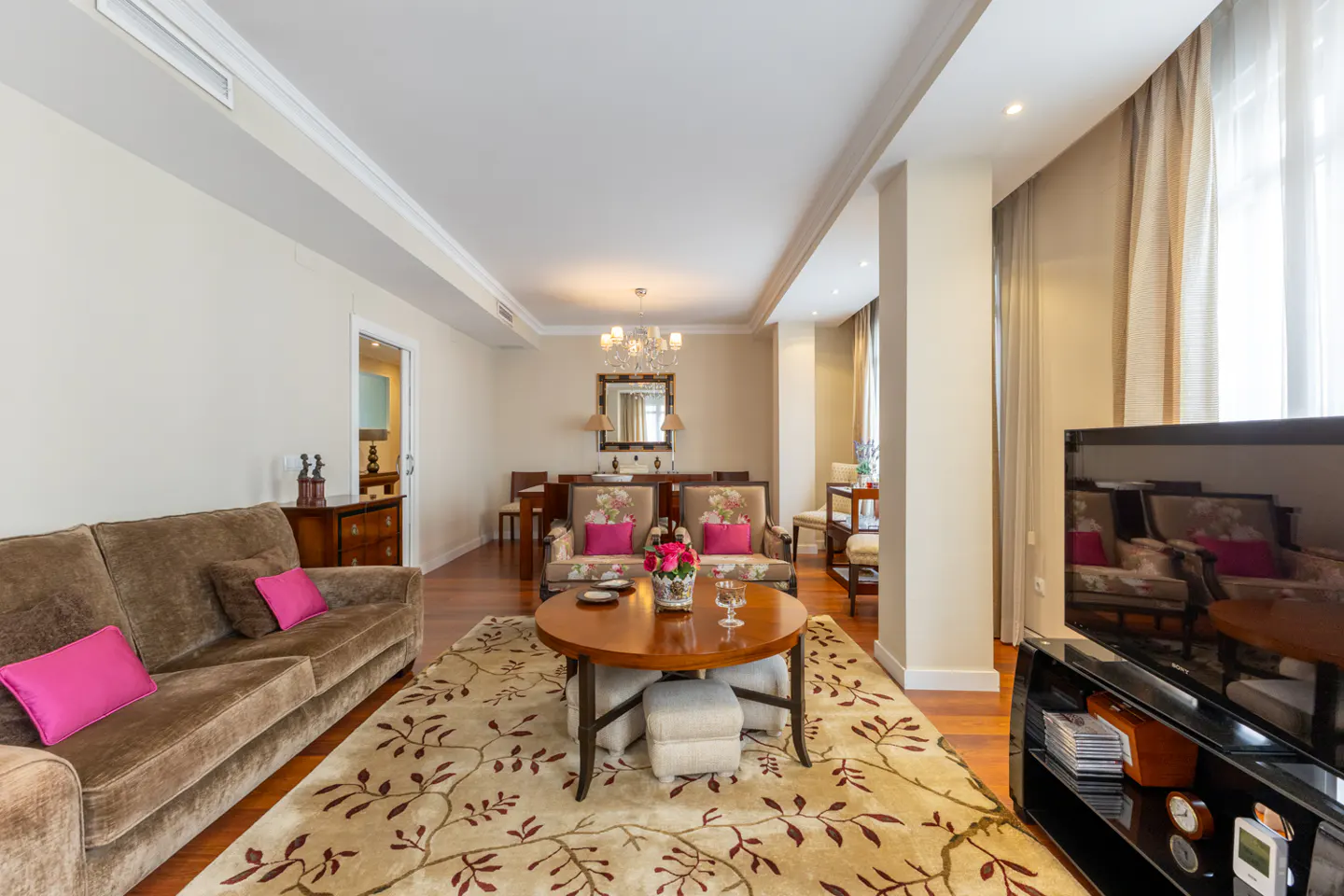 Elegant living room with a brown sofa, pink cushions, a round wooden coffee table, and a floral rug on hardwood floors.