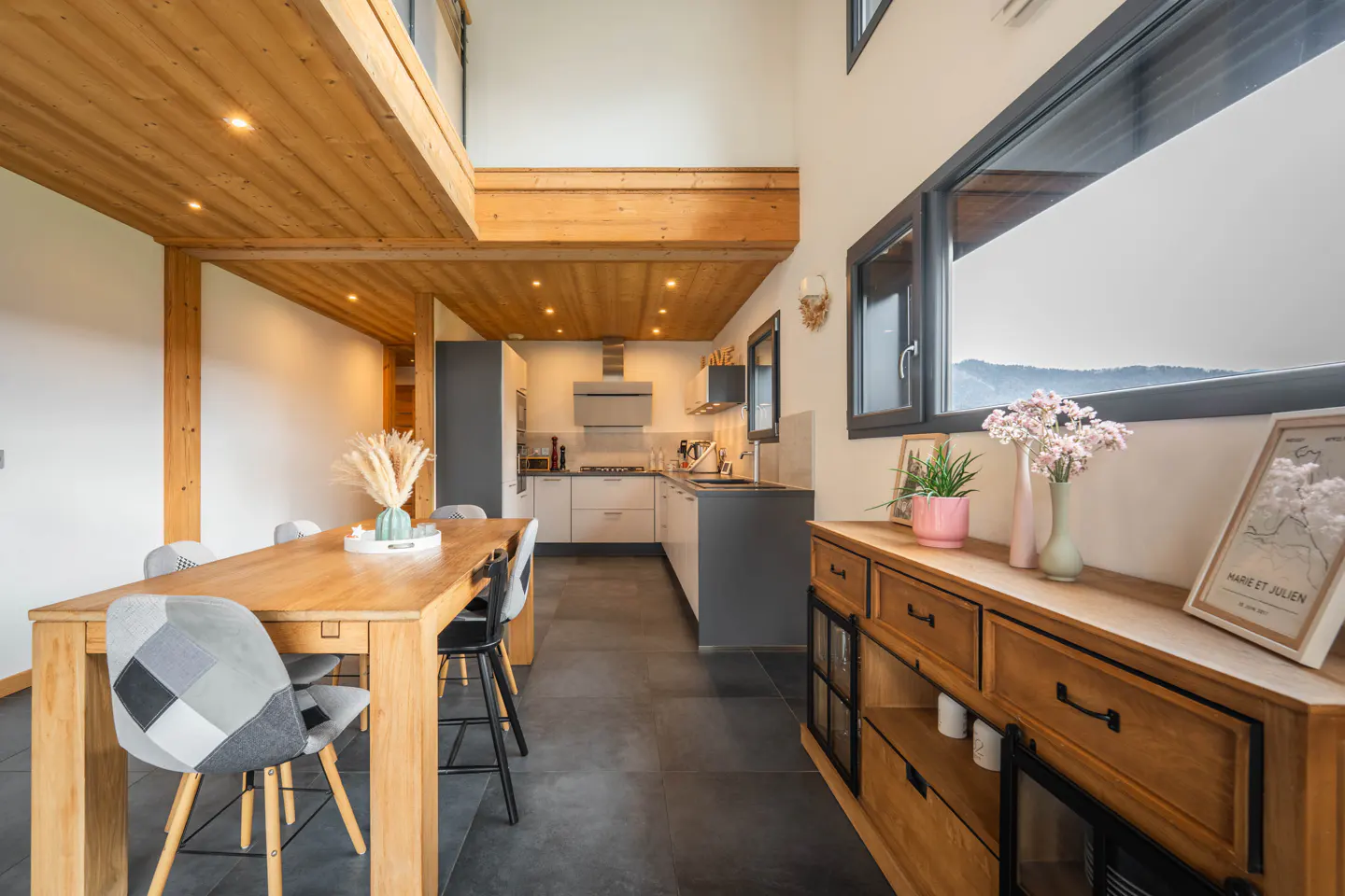 A modern kitchen and dining area with wood accents, a long table, and a large window with a mountain view.