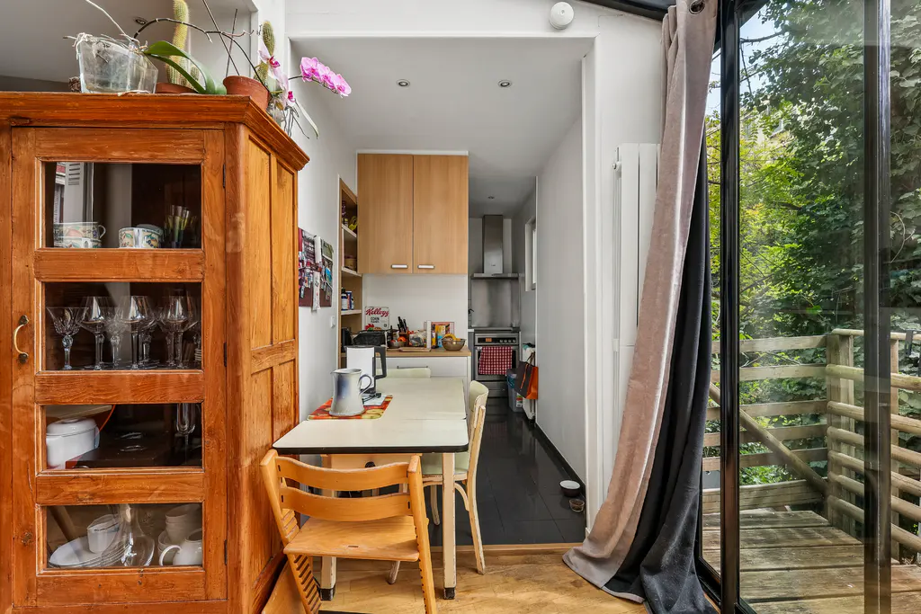 A bright kitchen with a wooden cabinet, table, and chairs. A glass door leads to a wooden deck and lush greenery.