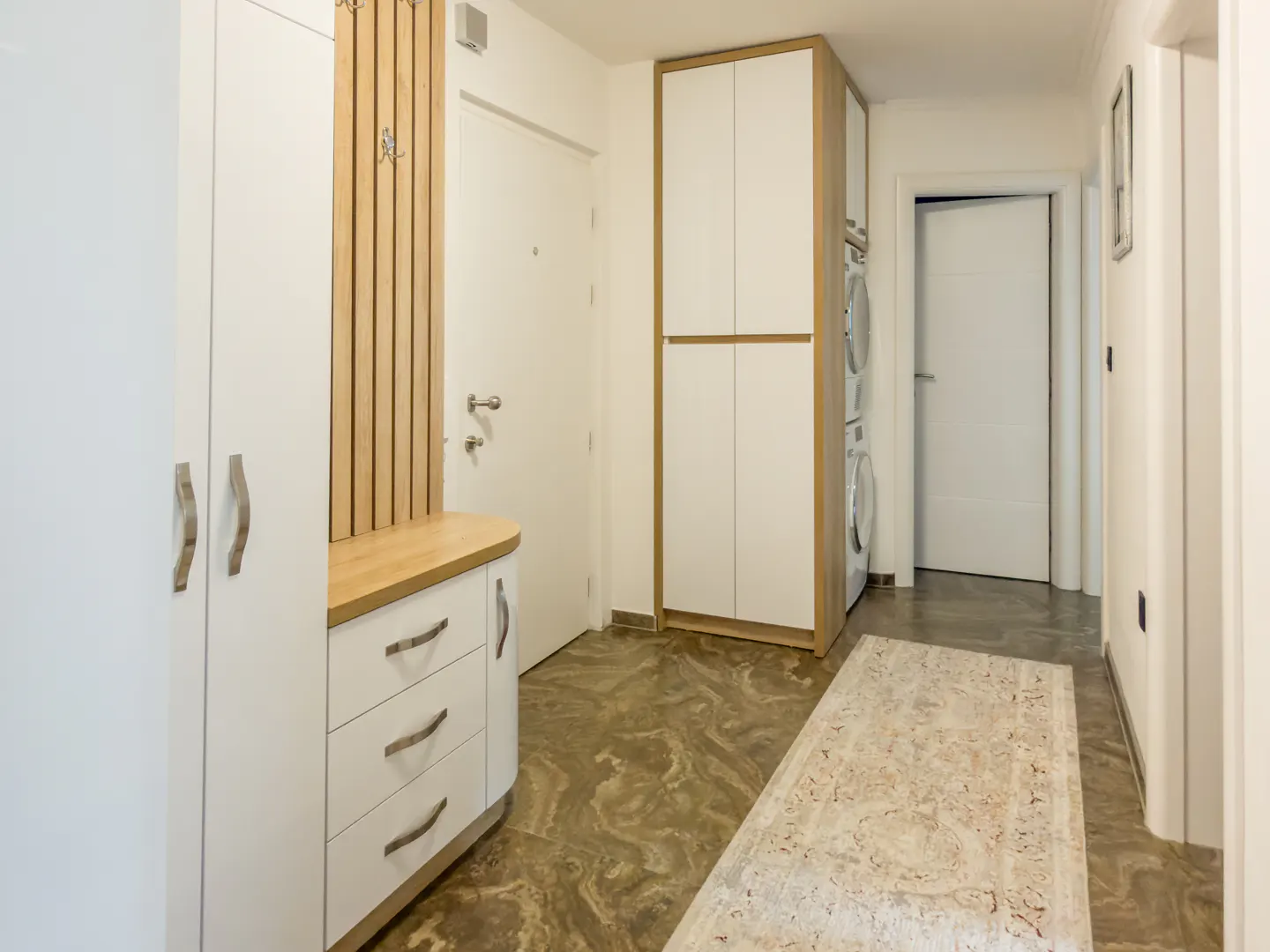 Hallway with white cabinets, drawers, and a coat rack. A beige rug lies on the brown marble floor. Washer and dryer are visible in the background.