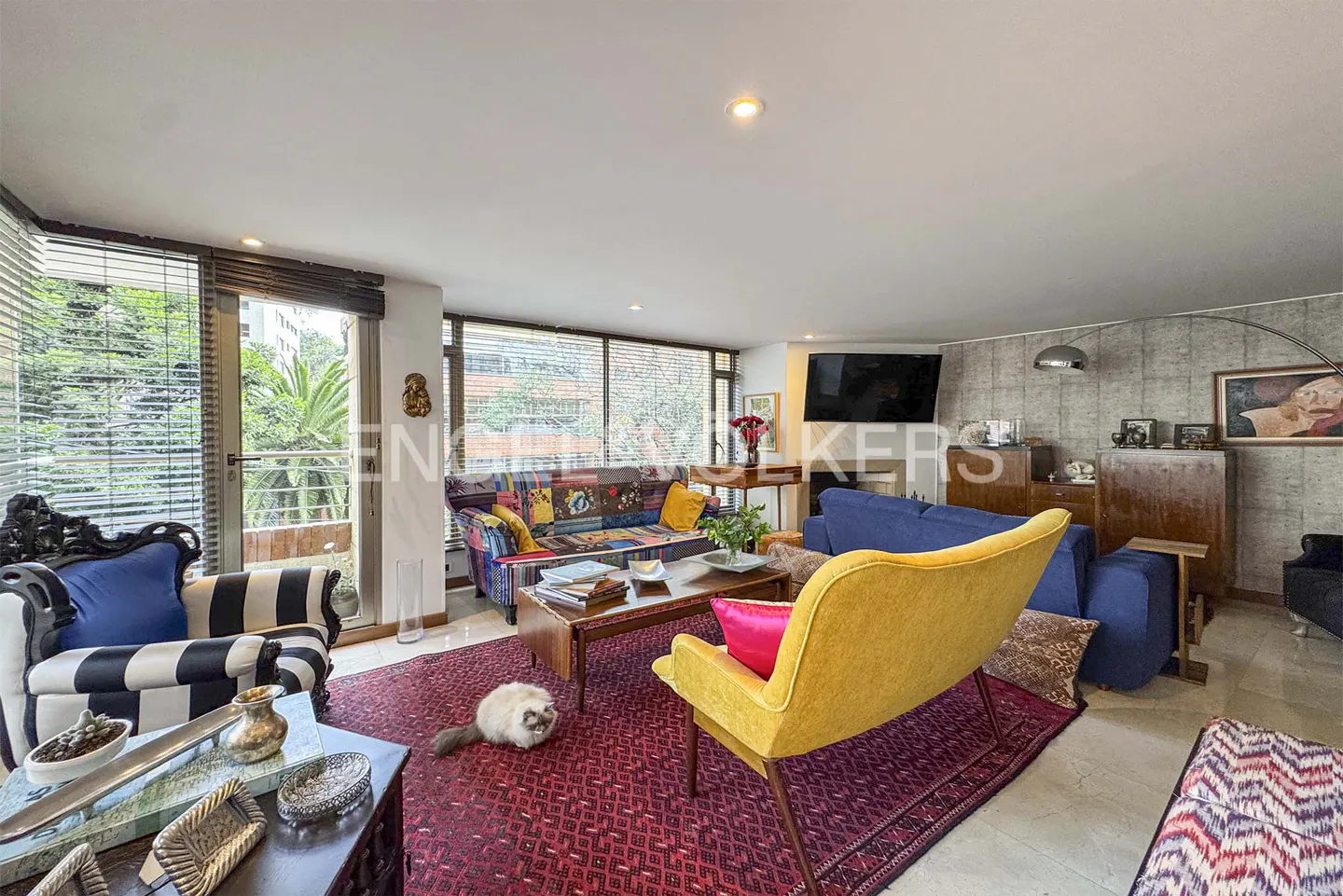 A living room with colorful furniture, a red rug, and a cat. Large windows offer a view of greenery.