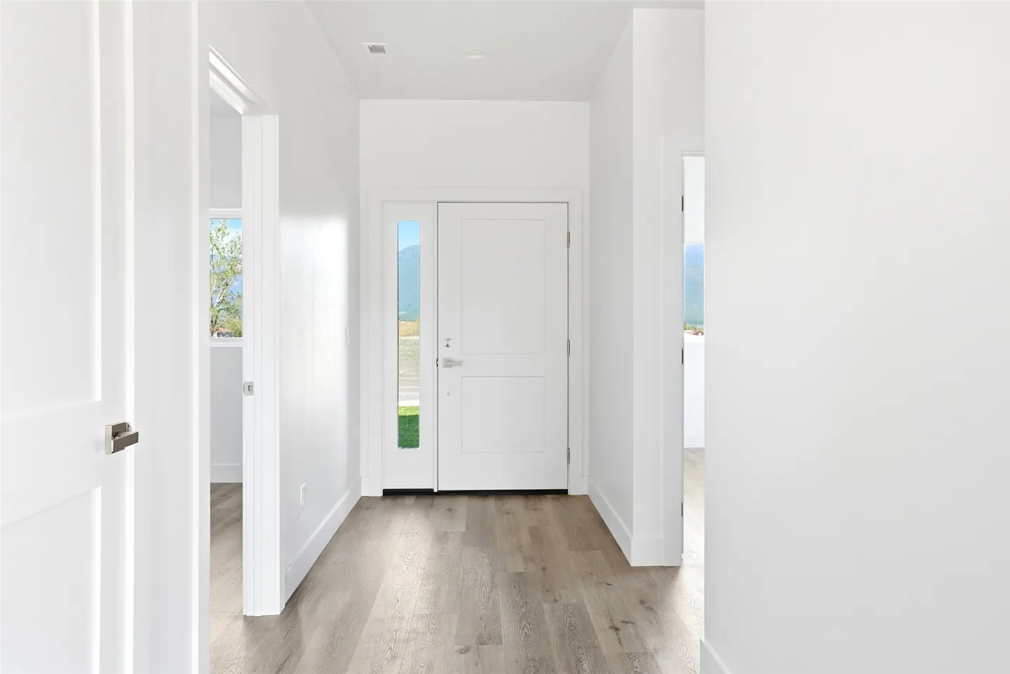 Bright, white entryway with wood-look floors. A white front door with a glass panel is centered. An open doorway shows a glimpse of a room.
