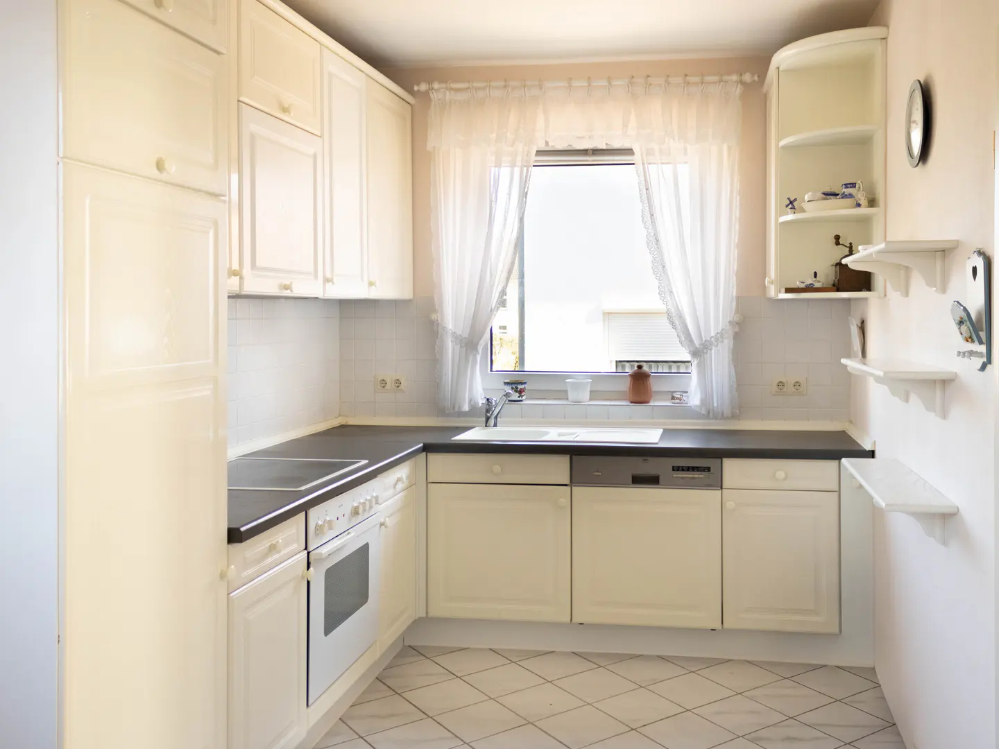 Bright kitchen with white cabinets, dark countertops, and a window with lace curtains. Shelves and a cabinet are on the right wall.