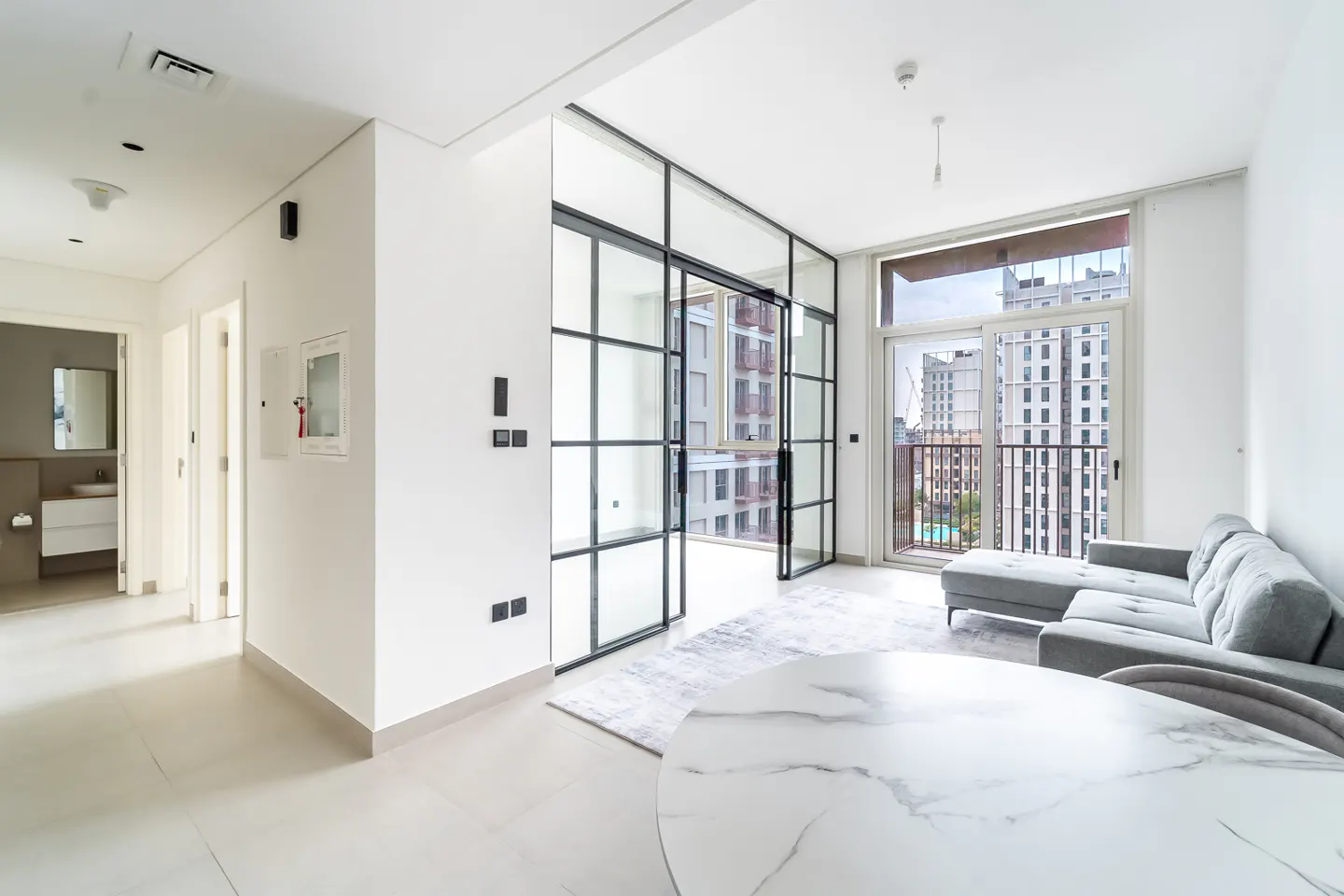 Bright, modern apartment interior with white walls, gray sofa, and marble table. Black-framed glass doors lead to a balcony with city views.