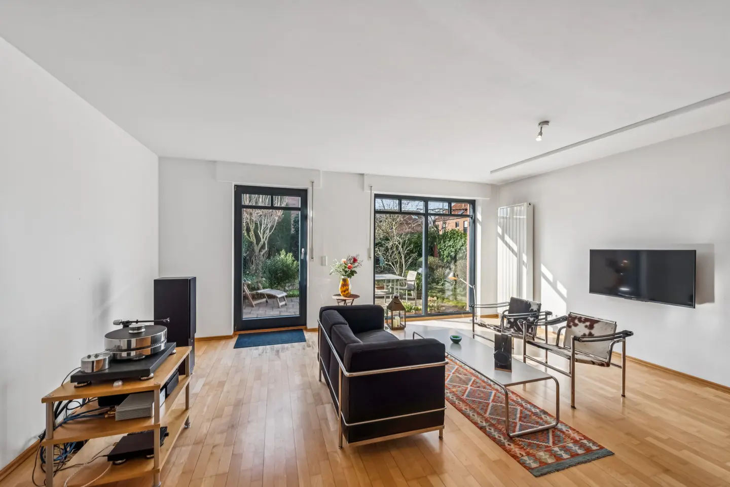 Bright living room with hardwood floors, black sofa, two chairs, and a TV. Doors lead to a garden with outdoor furniture.