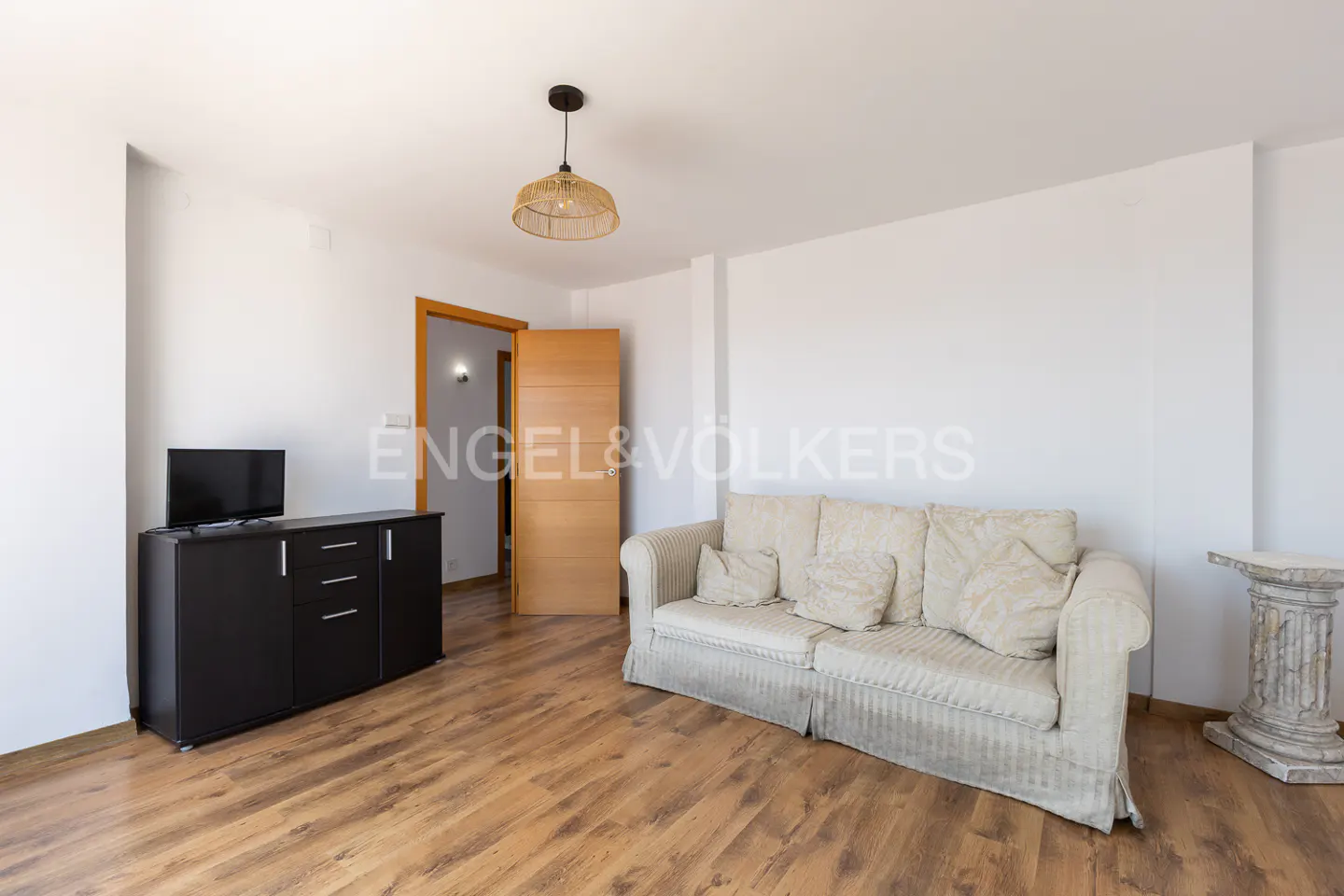 Bright living room with wood floors, white walls, beige sofa, black cabinet with TV, and open door.