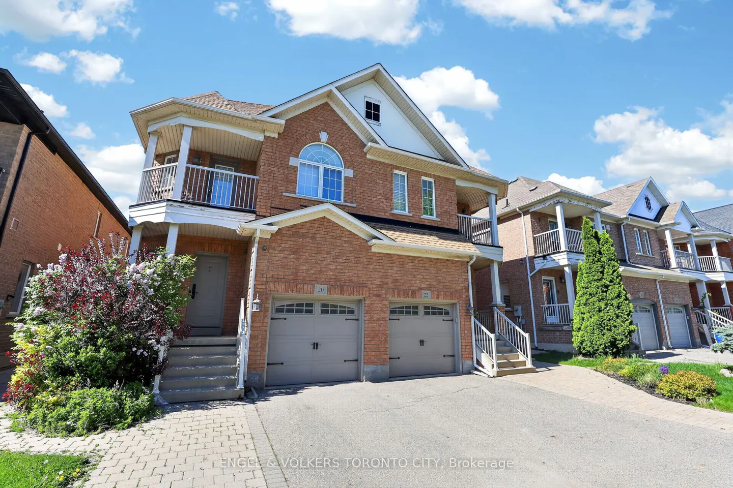 Two-story brick house with two-car garage, balconies, and a gray front door under a blue sky with white clouds.