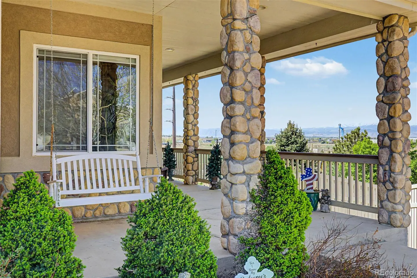 Covered porch with stone pillars, a white swing, and green bushes. A railing overlooks a landscape with trees and mountains in the distance.