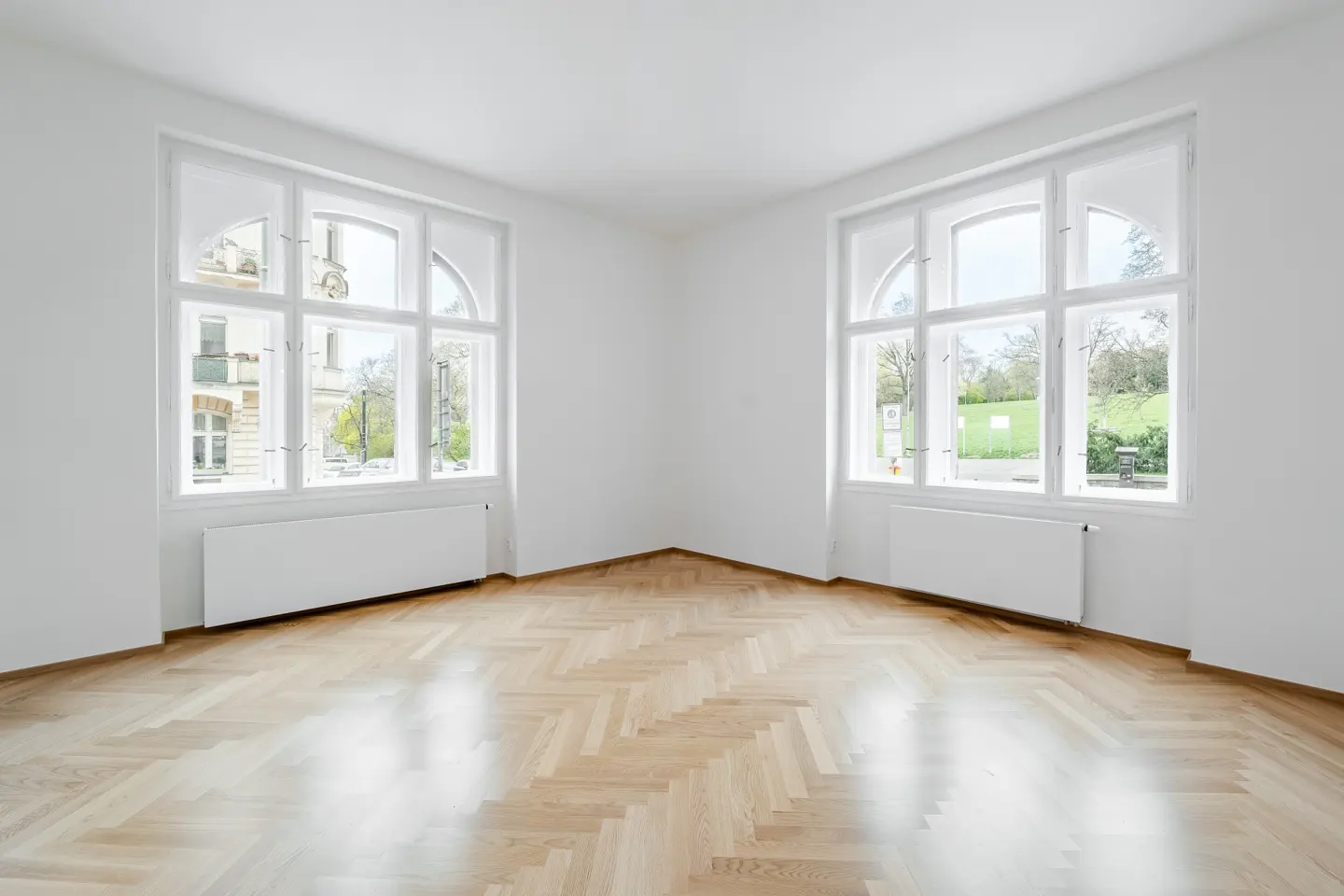 Bright, empty room with herringbone wood floors, white walls, and two large windows with views of buildings and greenery.