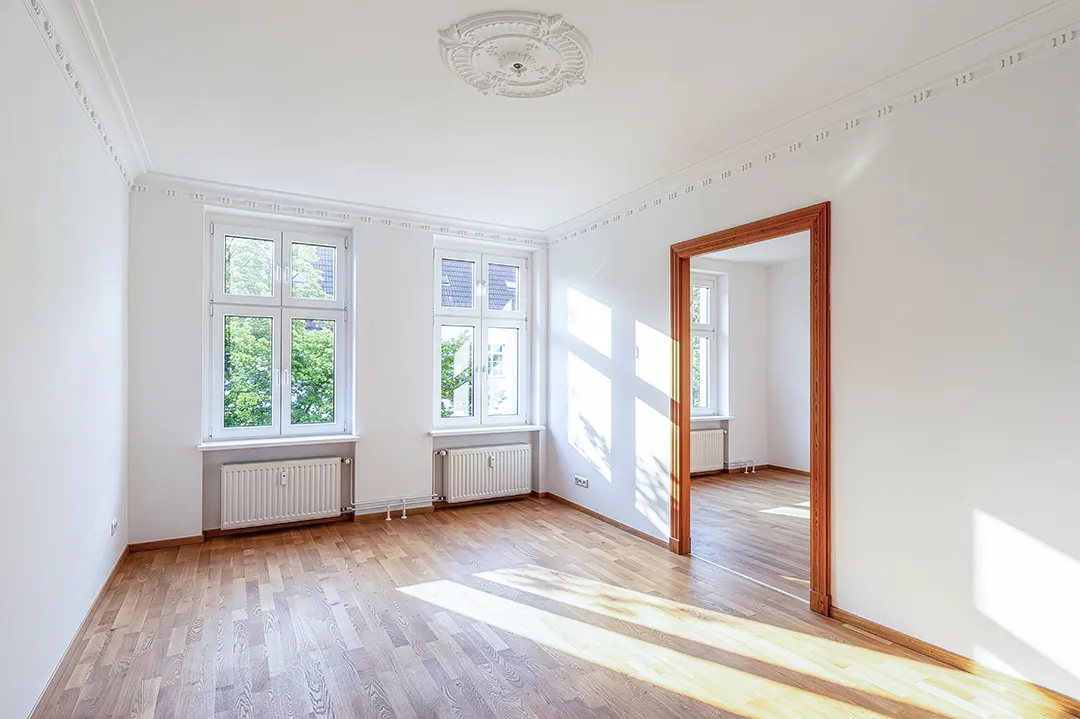 Bright, empty room with hardwood floors, white walls, and two windows. A doorway leads to another room with a window. Sunlight streams in.